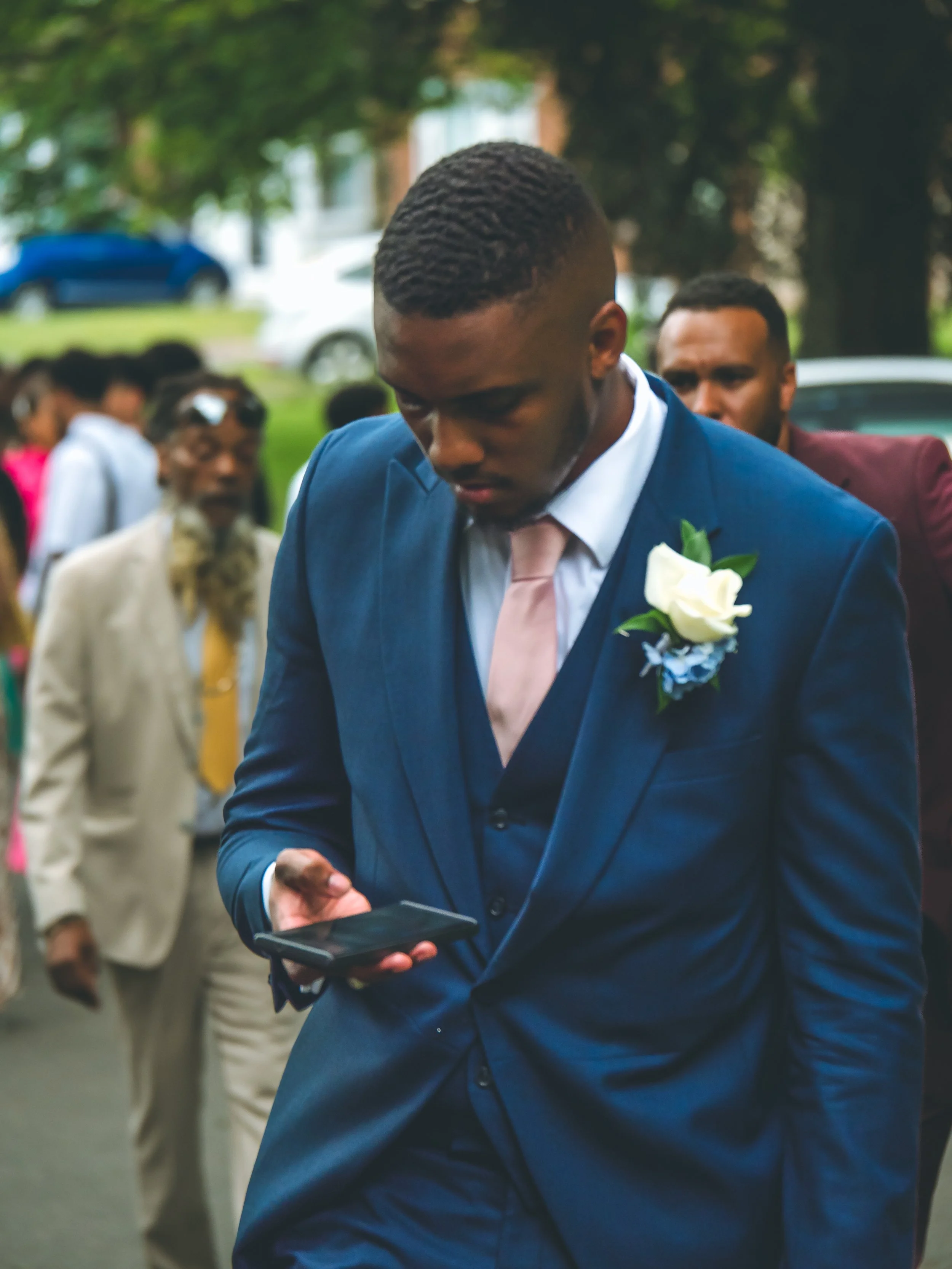 A man in a blue suit with a pink tie and white boutonniere looks down at his phone as he attends a formal outdoor event, with several people dressed in formal clothing and trees in the background.