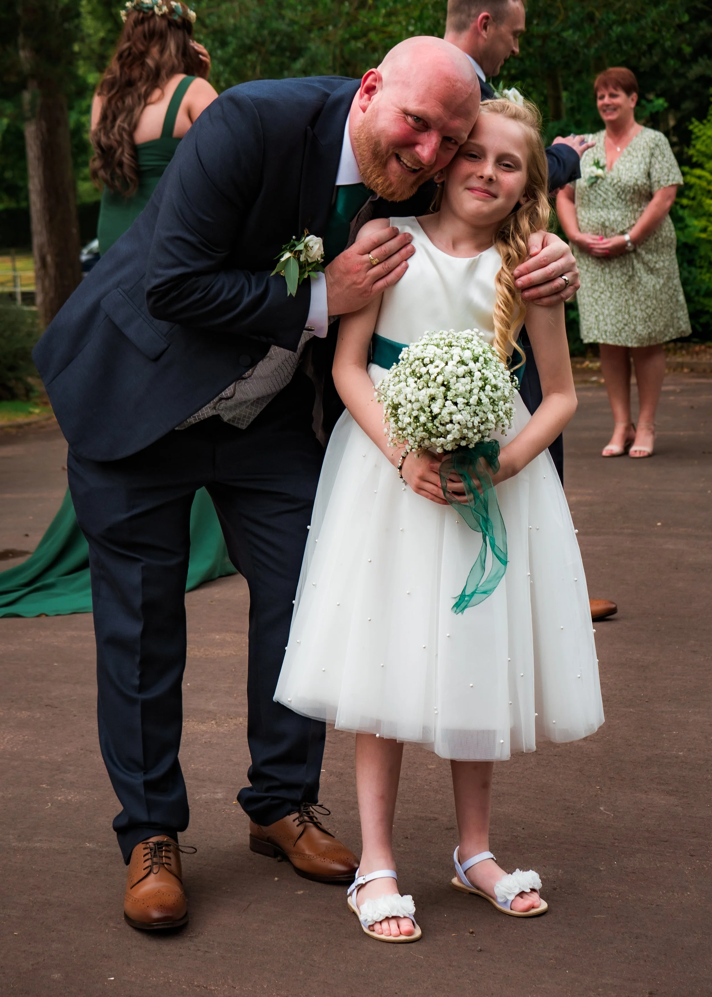 A group of people outdoors at a wedding, with a man and a young girl in the foreground. The man is leaning in and smiling, wearing a dark suit, and the girl in a white dress with green accents is holding a bouquet of small white flowers. In the backg