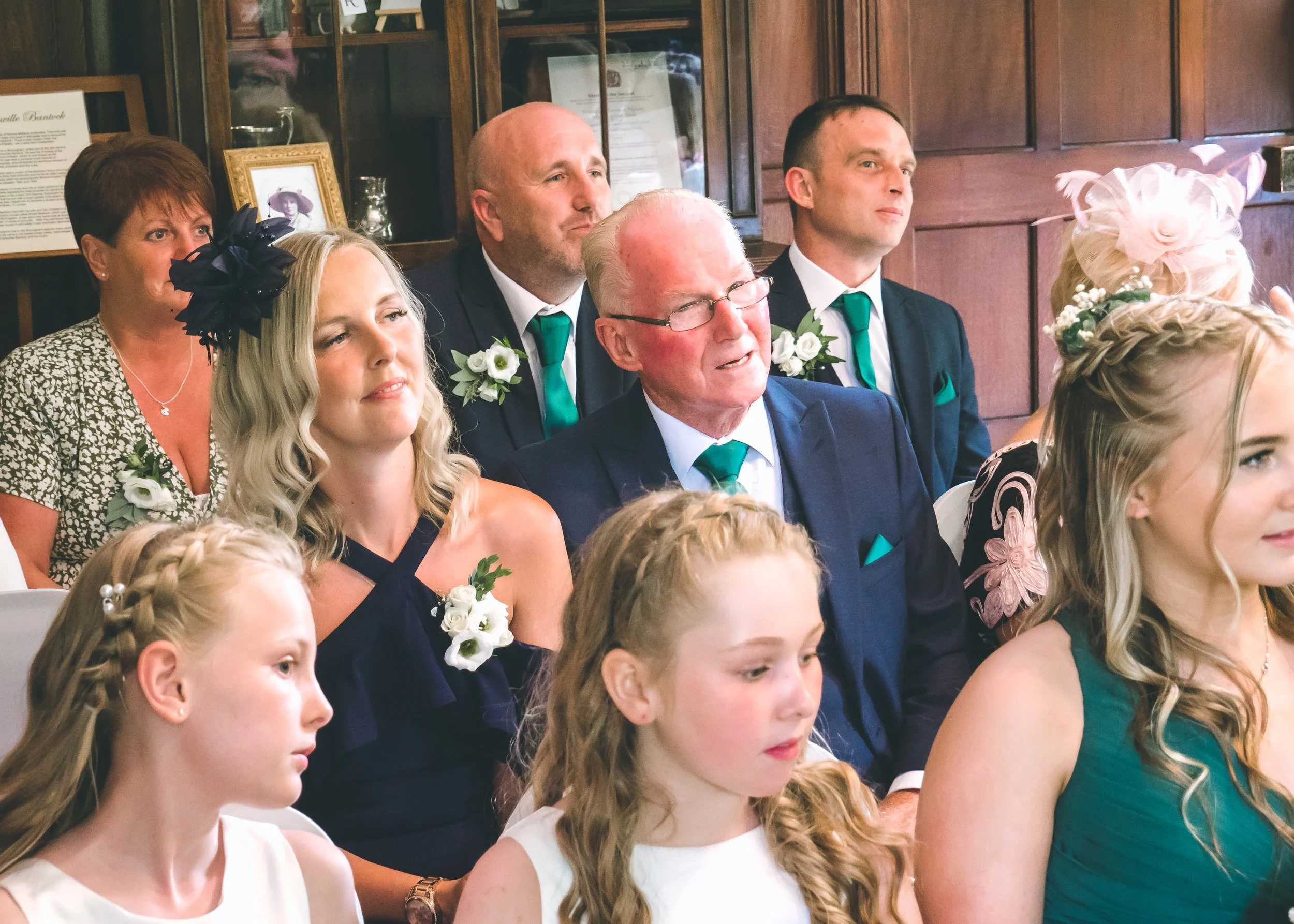 People dressed formally, sitting in a room with wooden walls, attending a celebration or ceremony. The women and men are wearing boutonnieres and ties, indicating a special occasion.