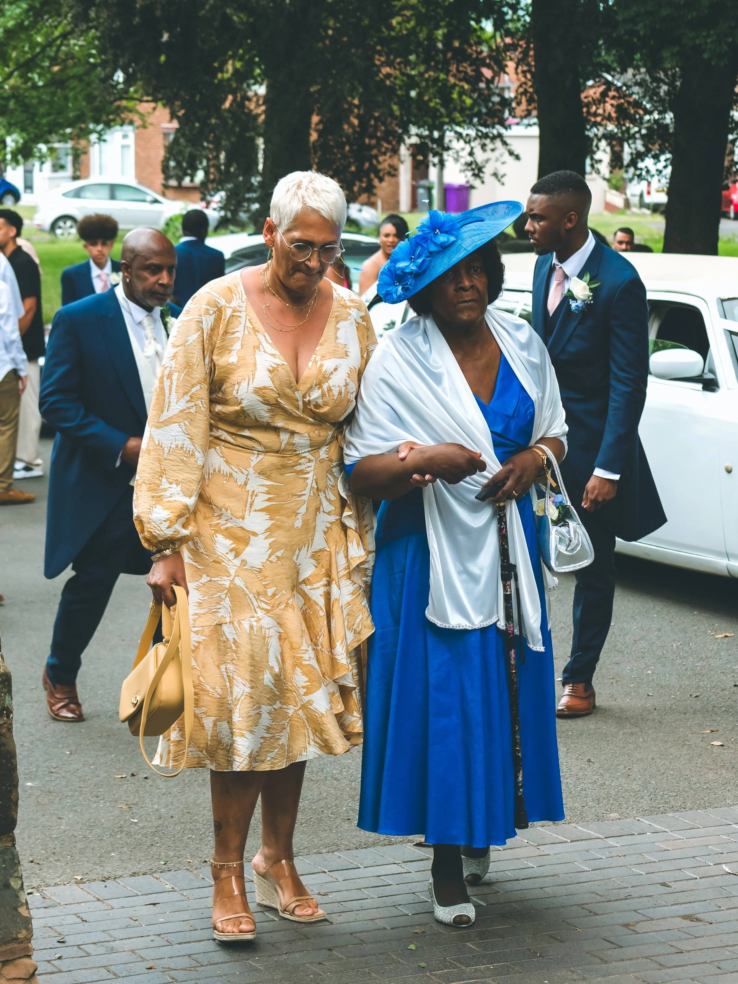 A group of people dressed formally walking outdoors, including women in dresses and men in suits, some wearing corsages and boutonnieres, likely attending a special event or celebration.