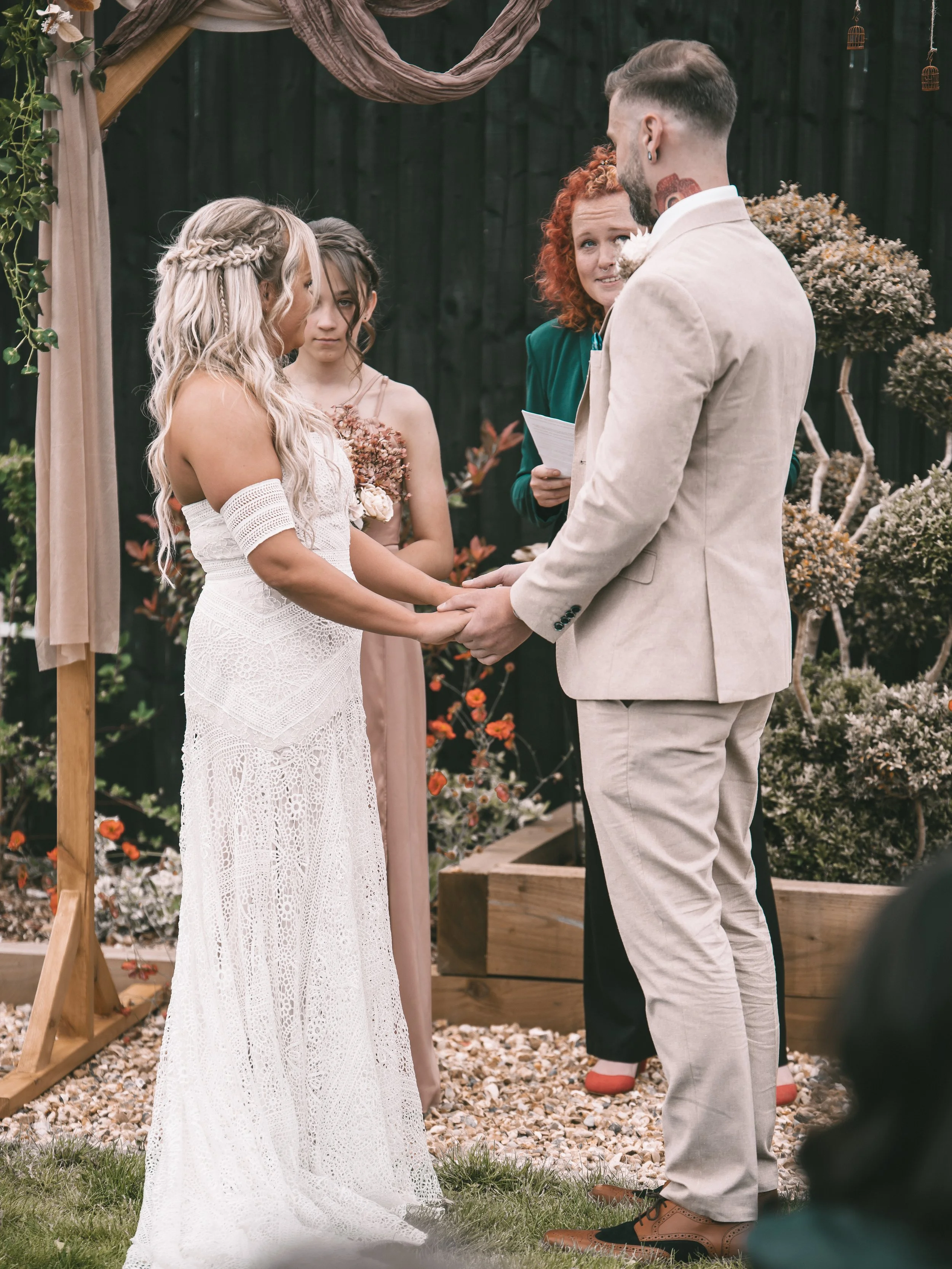 A couple getting married outdoors, holding hands, during their wedding ceremony. The bride is wearing a white lace dress with an off-shoulder design and braided hair. The groom is dressed in a light-colored suit with brown shoes. An officiant is stan