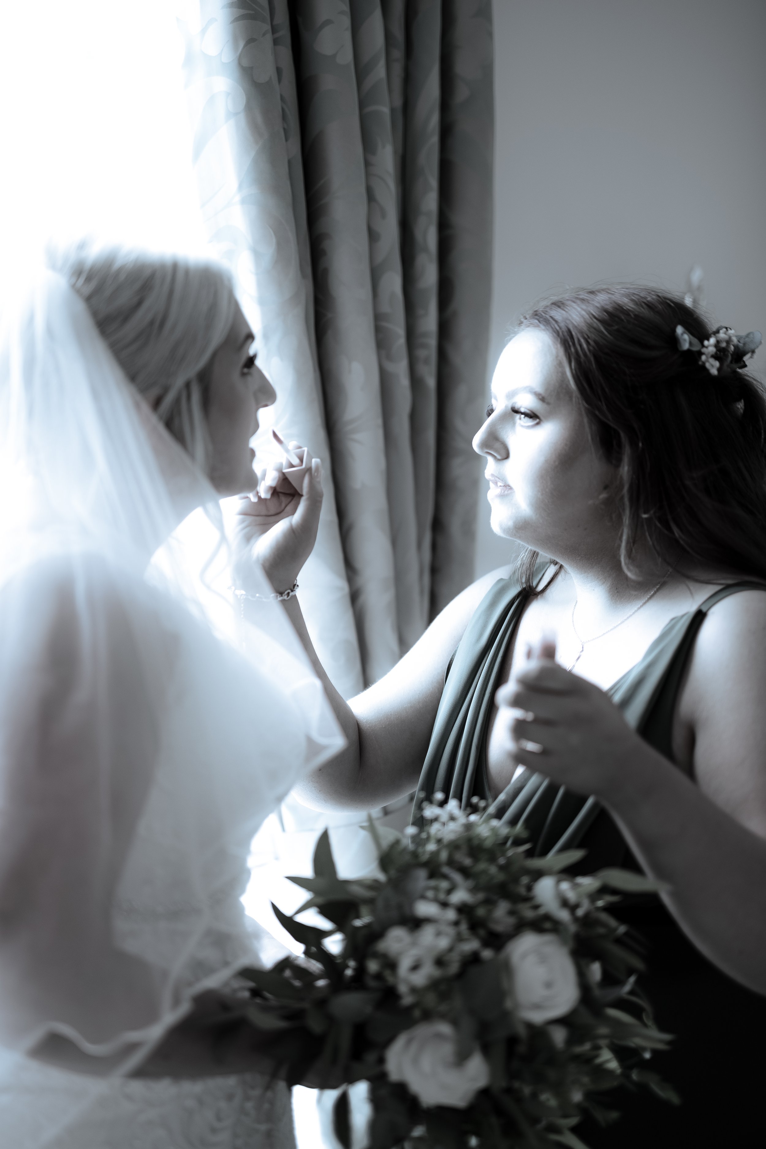 Two women, one in a wedding dress and veil, and the other with long hair adorned with flowers, face each other near a window with curtains, exchanging a tender moment.