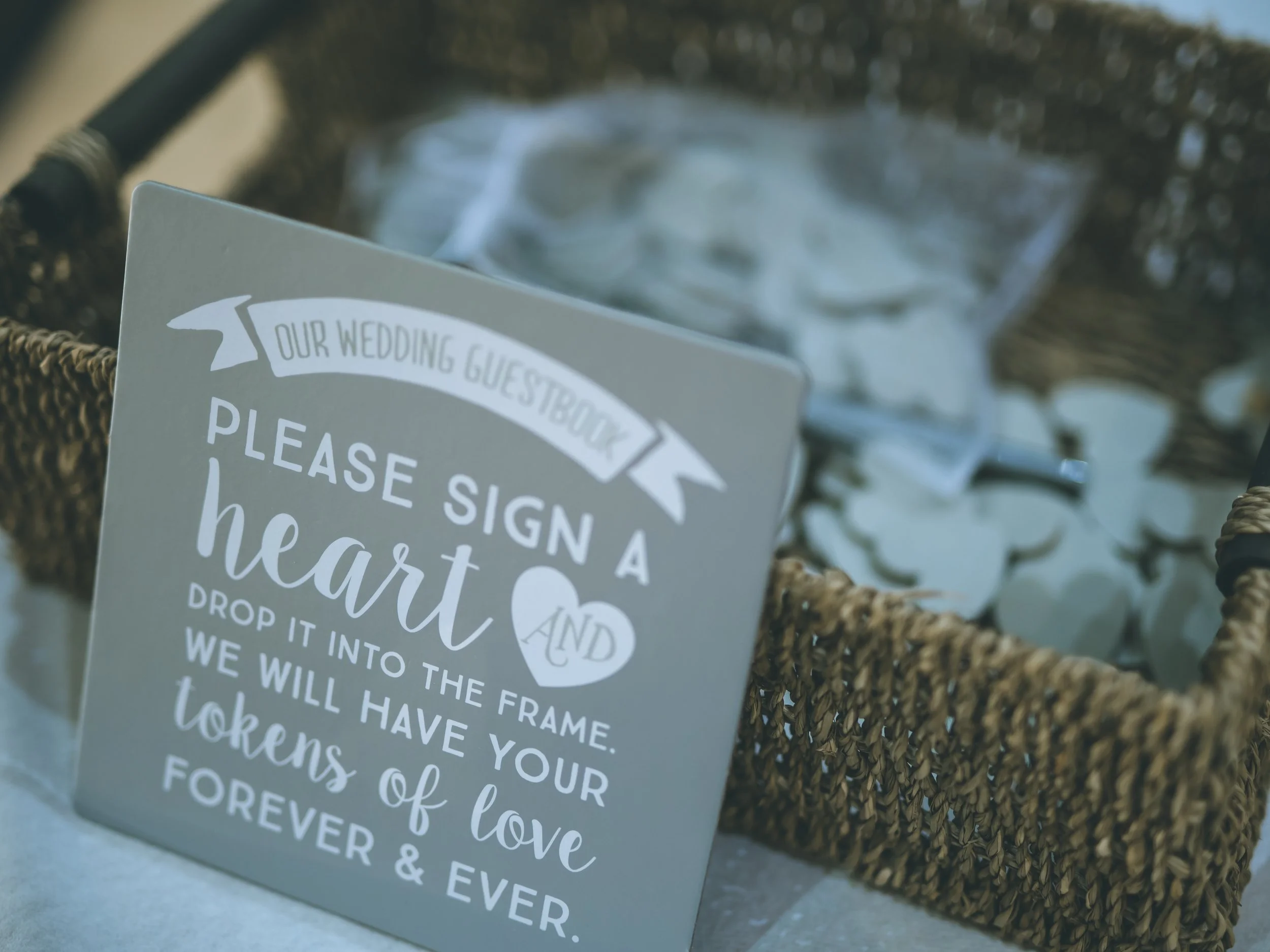 A printed sign in a basket with a blurred background of trees and sky. The sign appears to have a heartfelt message with the words "please sign a heart" and other text about love and memories.