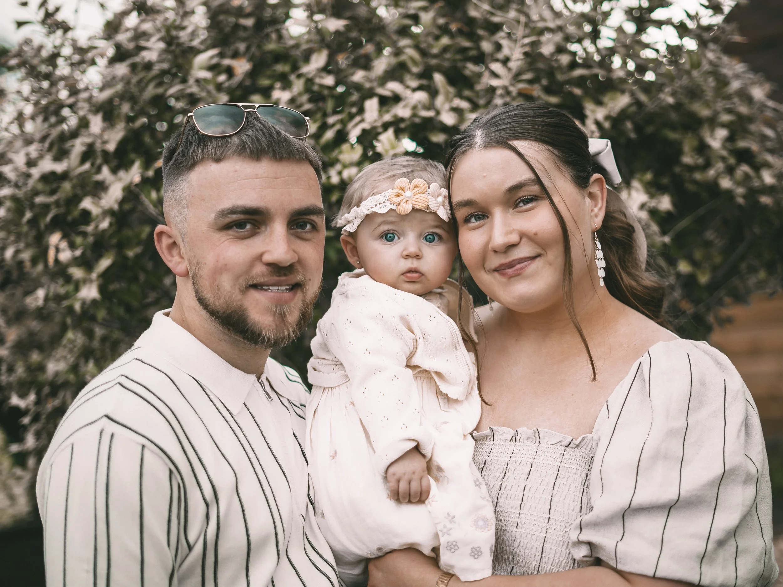 A family of three, a man, a woman, and a baby, standing outside in front of green bushes. The man has sunglasses on his head and wears a white shirt with black stripes. The woman has long dark hair tied back with a white ribbon and is smiling. The ba