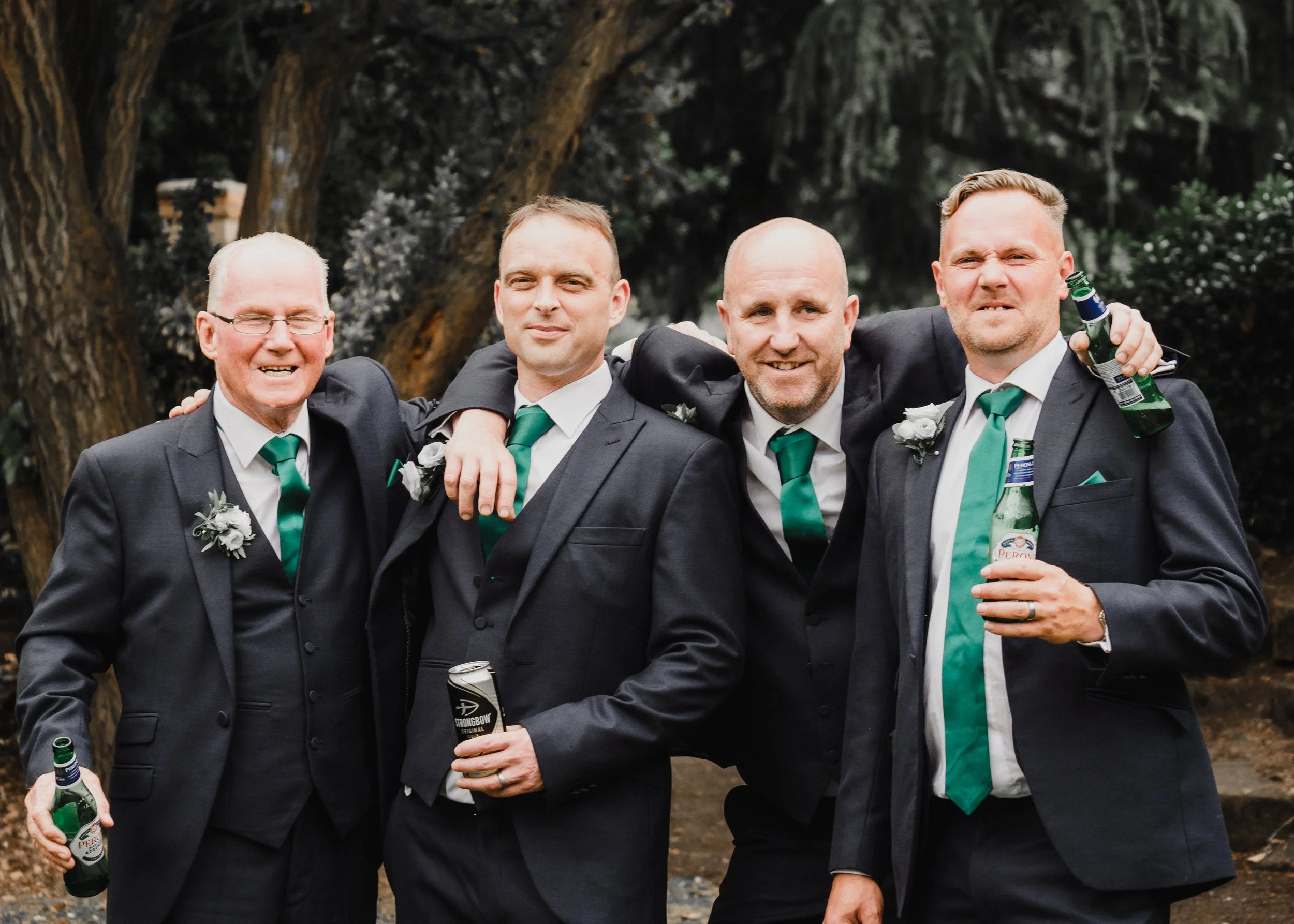 Four men in suits with white shirts and green ties standing outdoors. They are smiling and have their arms around each other. They are holding green bottles of beer, and they appear to be celebrating at a formal event or gathering.