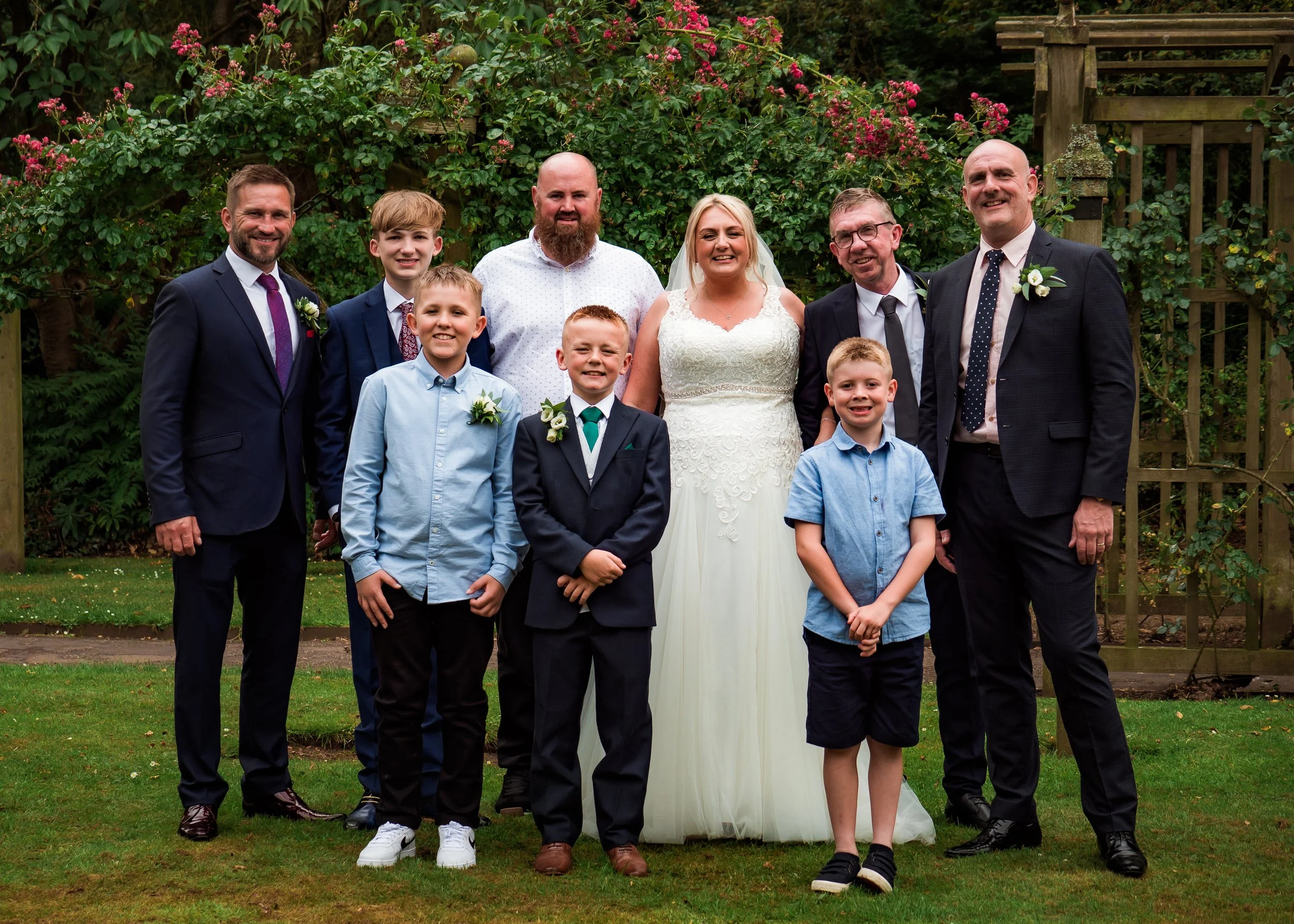 A group of nine people, including an bride in a white wedding dress, posing outdoors in front of green bushes with pink flowers. They are dressed formally, with some men in suits and ties, and children in dress shirts. The group is smiling and standi