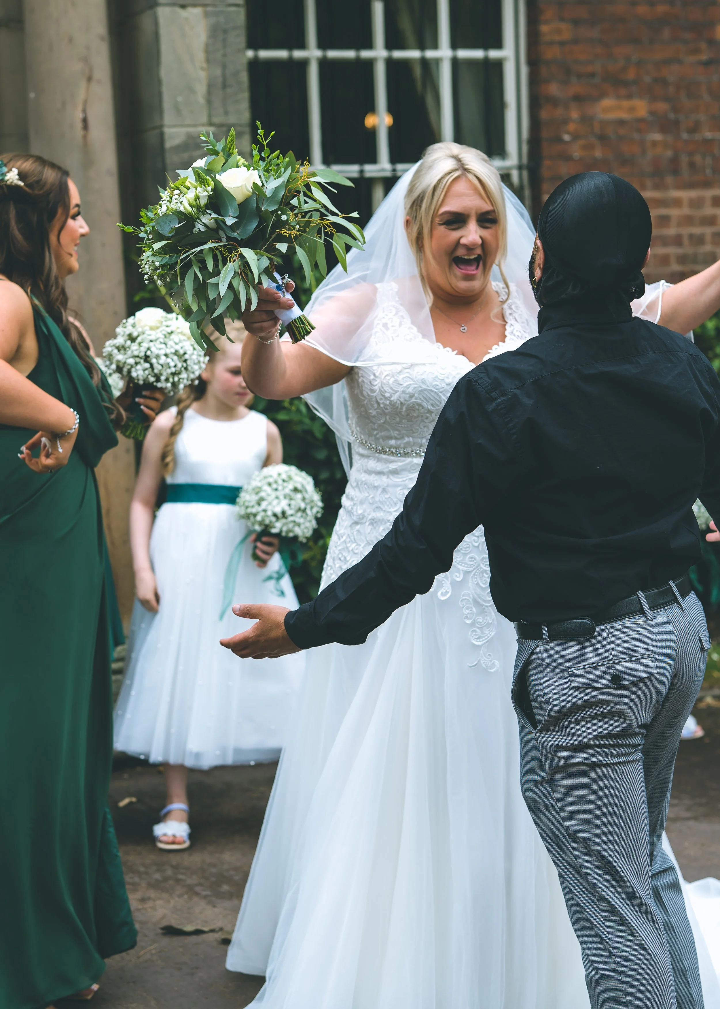 A bride in a white wedding dress and veil excitedly celebrates with a groom in gray checked pants and a black shirt at her wedding. A bridesmaid in a green dress and two young girls in white dresses hold bouquets in the background, outside a brick bu