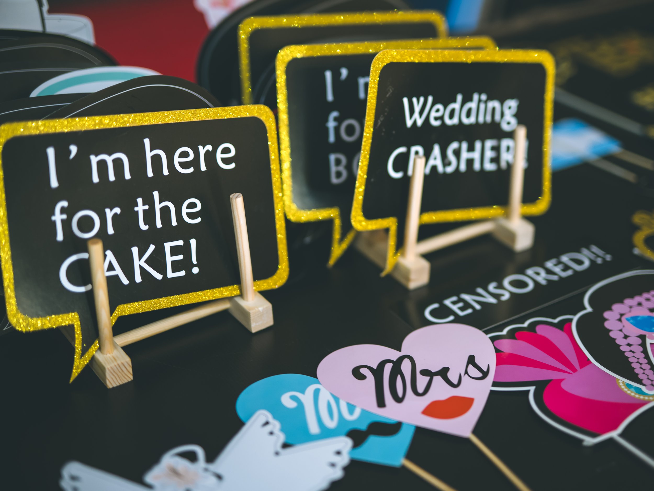 Colorful decorative signs with messages for special occasions displayed on small wooden stands. The signs have black backgrounds with glittery gold borders and white or colorful text, including phrases like "I'm here for the cake!" and "Wedding cashi