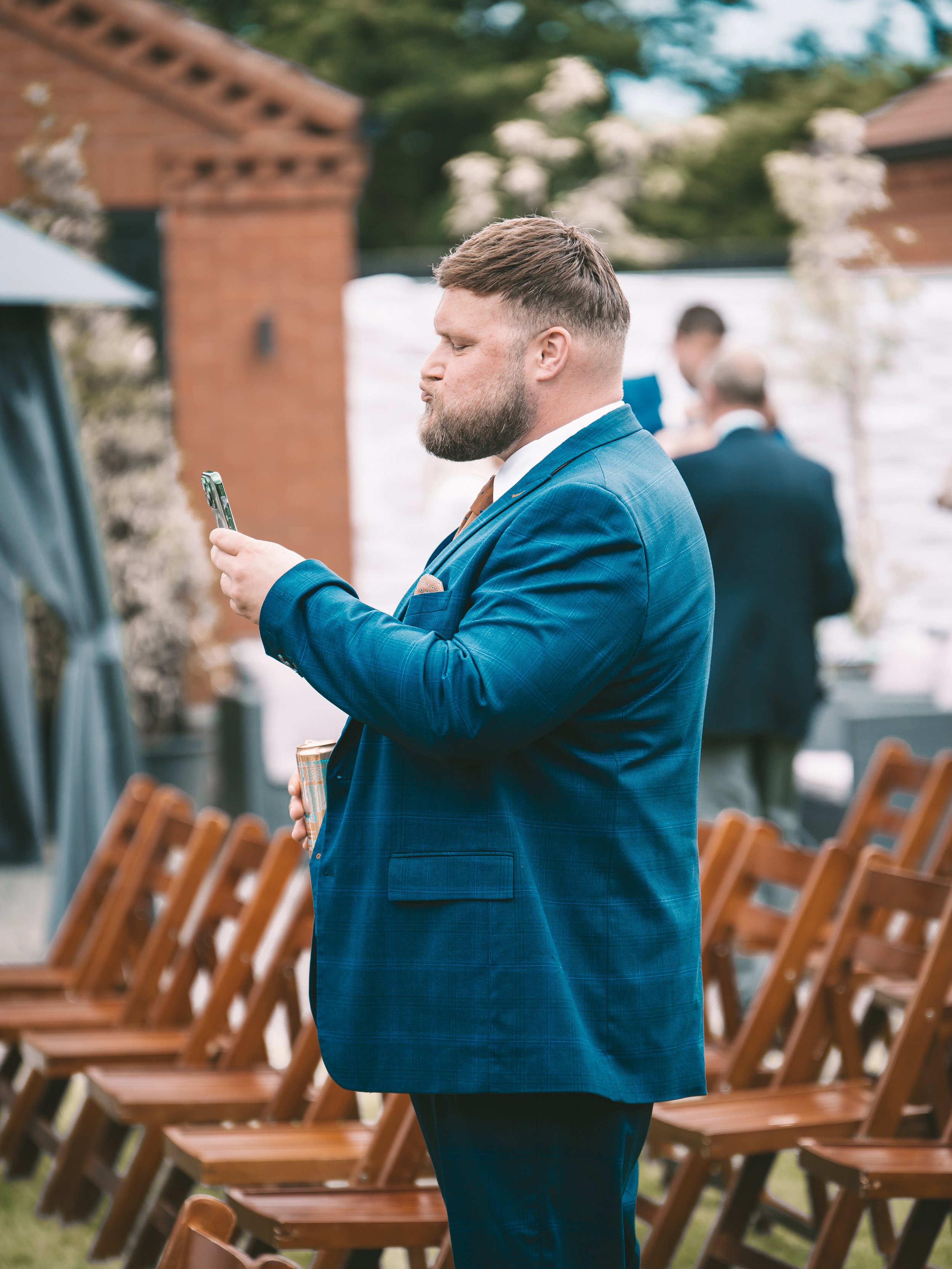 A man with a beard in a blue suit jacket practicing a seated yoga pose outdoors at a wedding or event, with rows of wooden chairs and other people in the background.