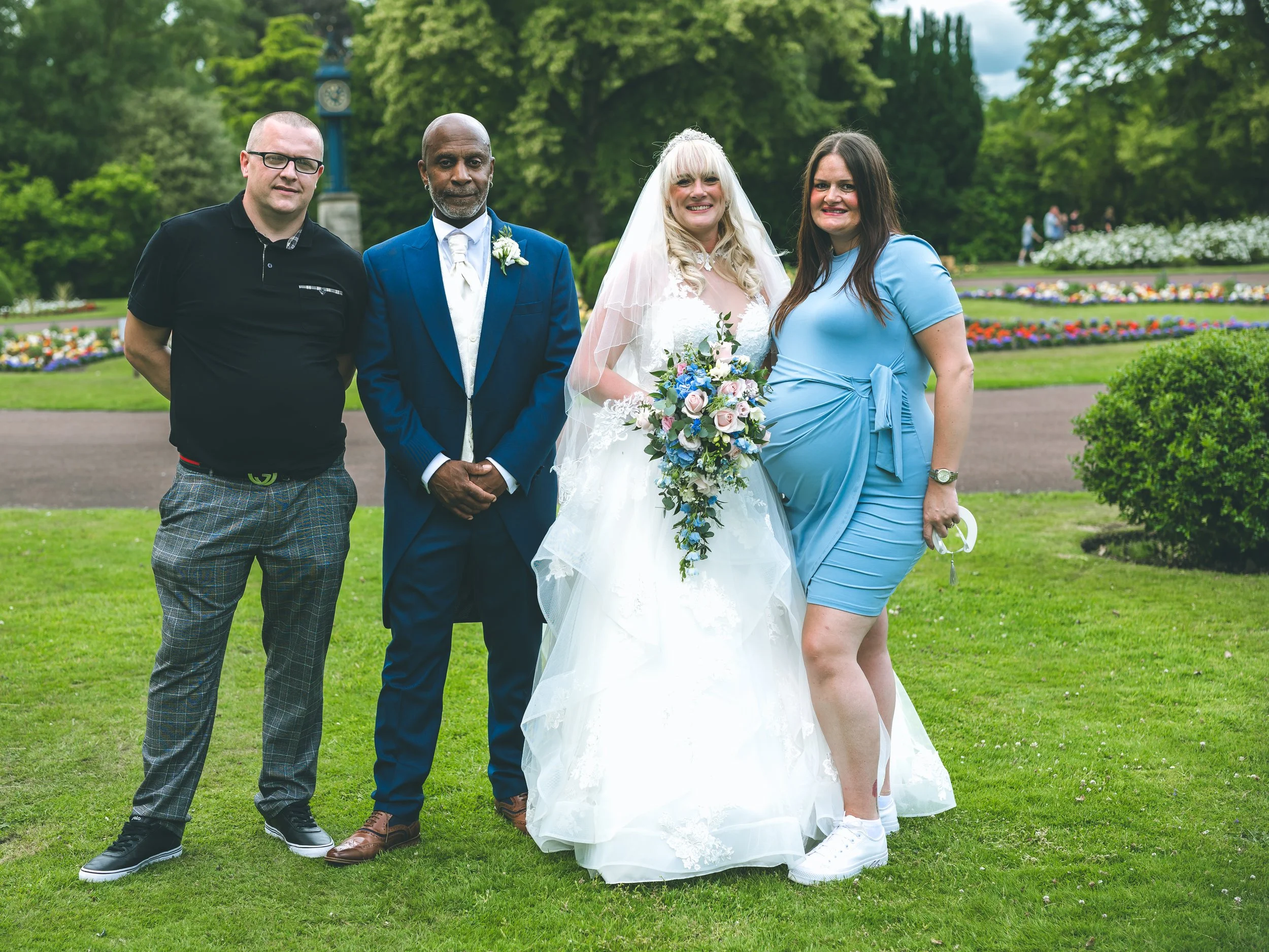 Group of four people standing on grass in a park with colorful flowers and green trees in the background. The group appears to be celebrating a wedding, with the woman in a white wedding dress holding a bouquet and wearing a veil. The others are casu