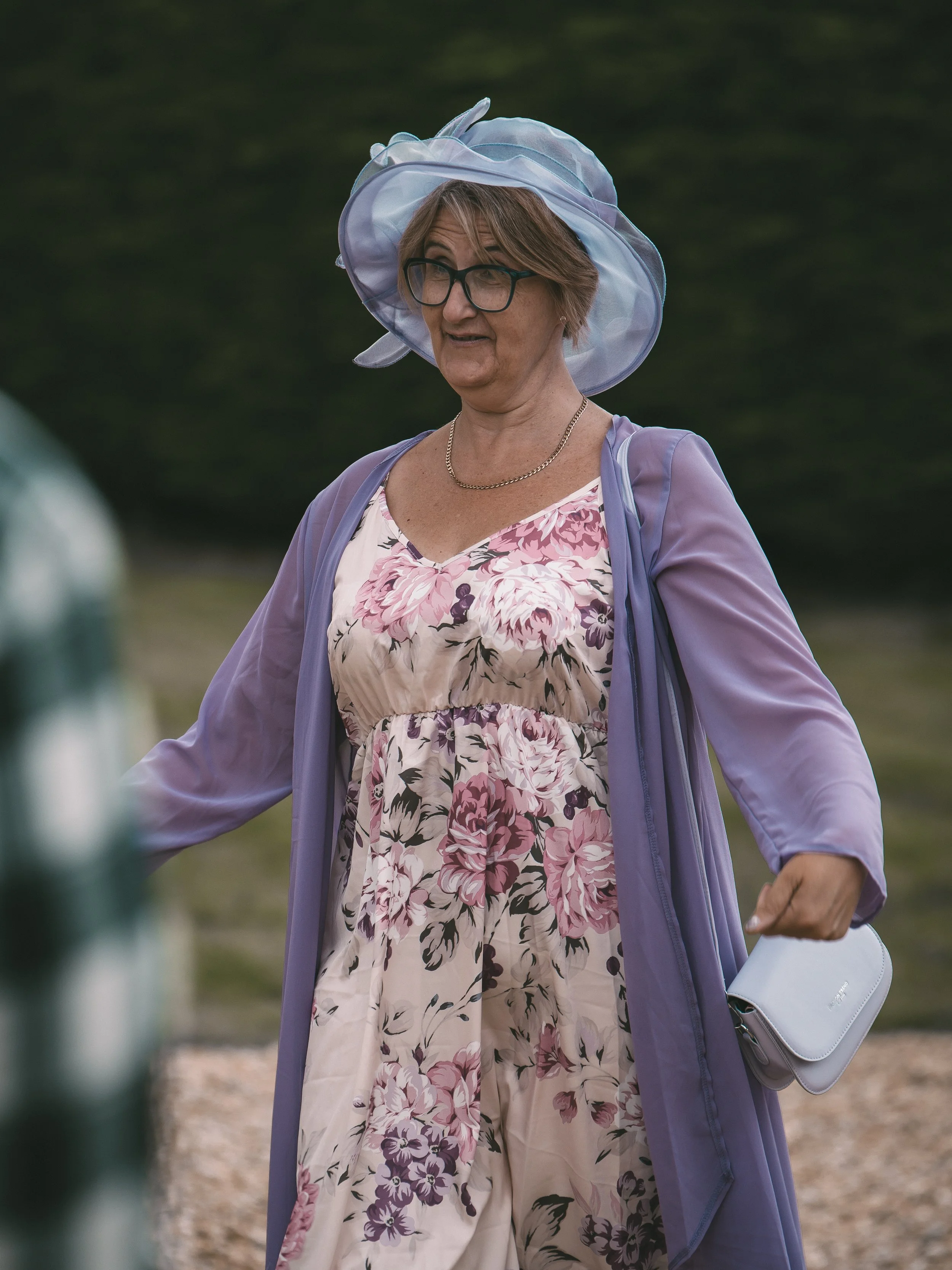 An older woman wearing a large blue hat, glasses, a purple cardigan, and a floral dress, carrying a gray purse and walking outdoors.