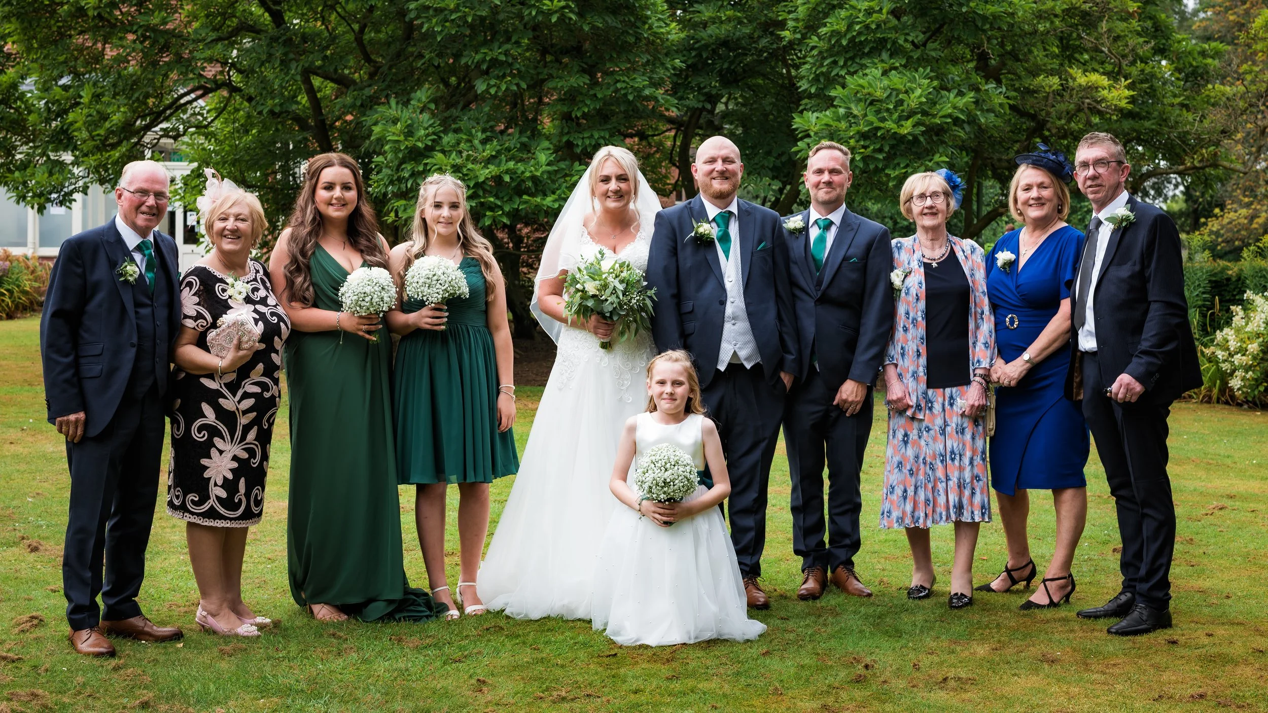 Group of people at a wedding outdoors, including bride, groom, children, and family members, standing on a grassy area with trees in the background.