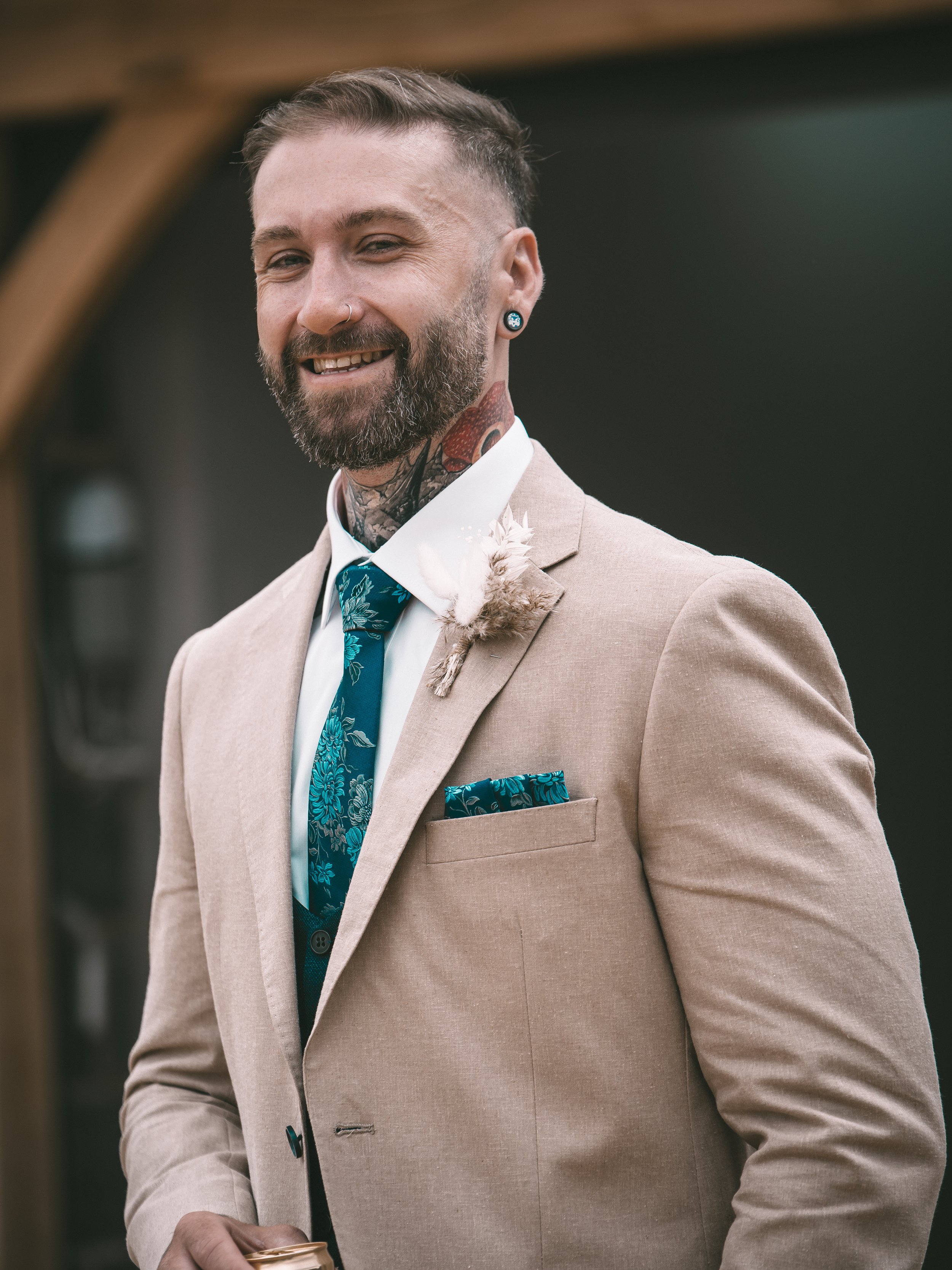 A man with a beard and tattoos wearing a beige suit, white shirt, teal tie, and a boutonniere, smiling and holding a drink, standing outdoors.