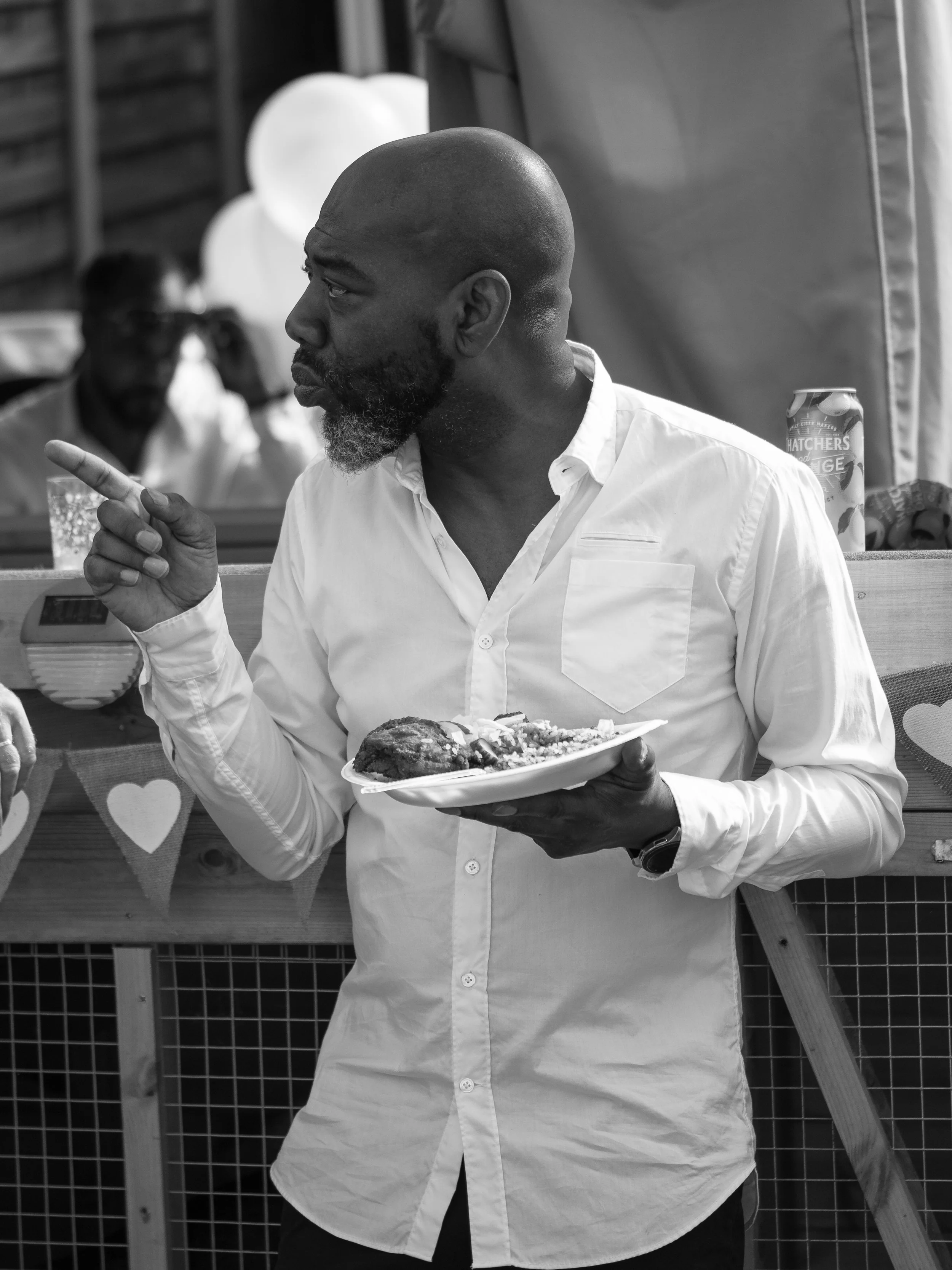 A man with a bald head and beard wearing a white shirt, holding a plate of food, pointing with his finger, at an outdoor event.