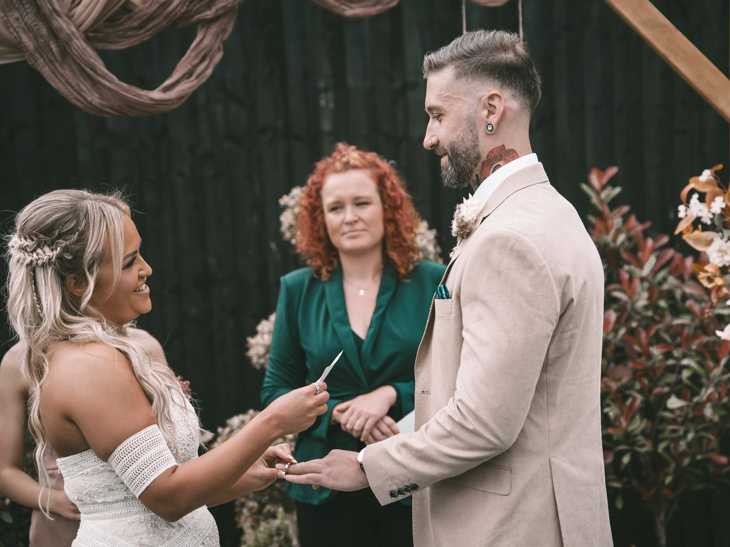 A couple exchanging wedding rings during a ceremony, with a woman officiant and a man in a beige suit, set outdoors with floral decorations.