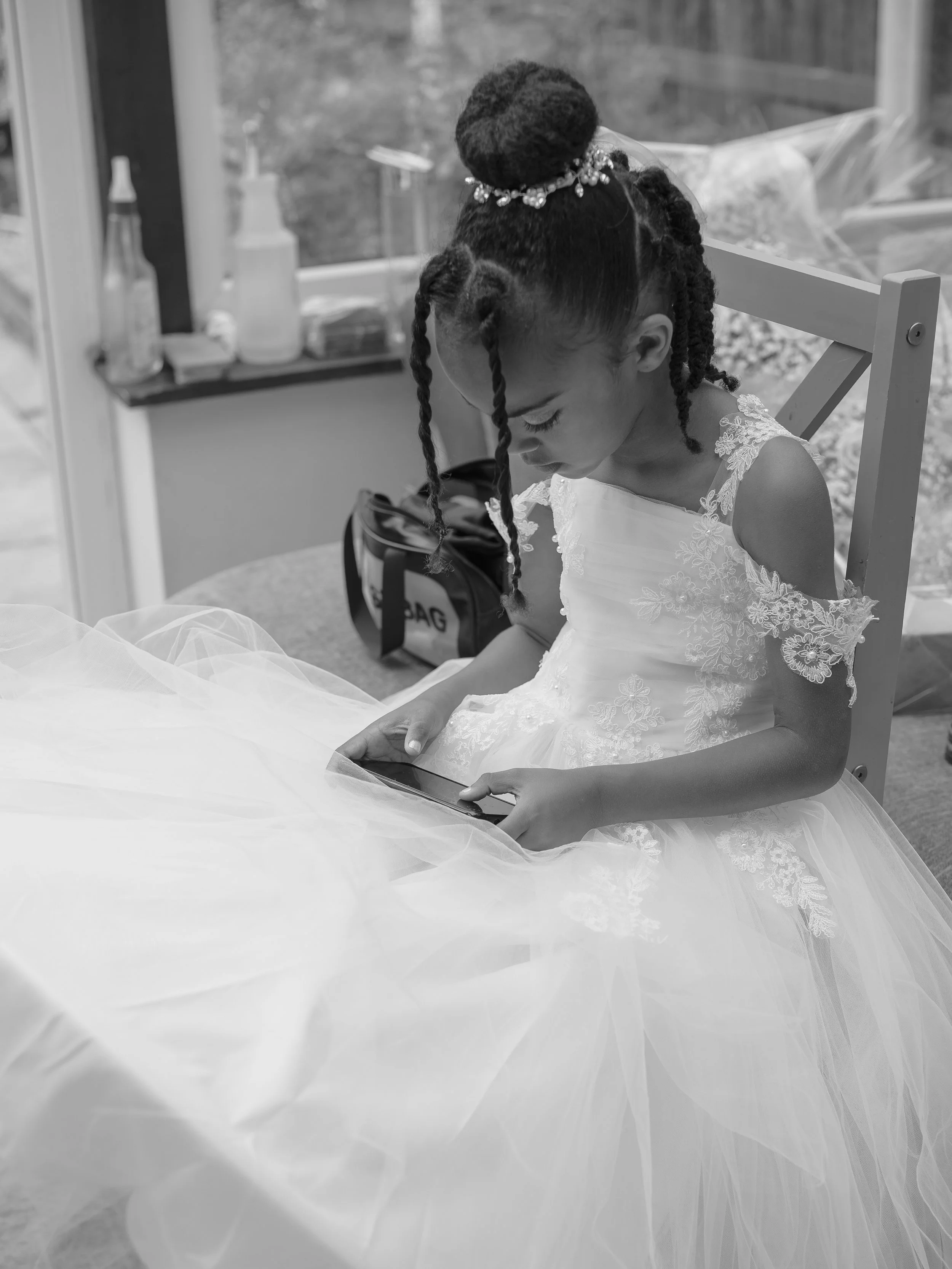 A young girl in a white lace dress with floral embroidery is sitting at a table, looking down at a smartphone, with an elegant braided hairstyle and a hair accessory, in a room with a window and various bottles and objects in the background.