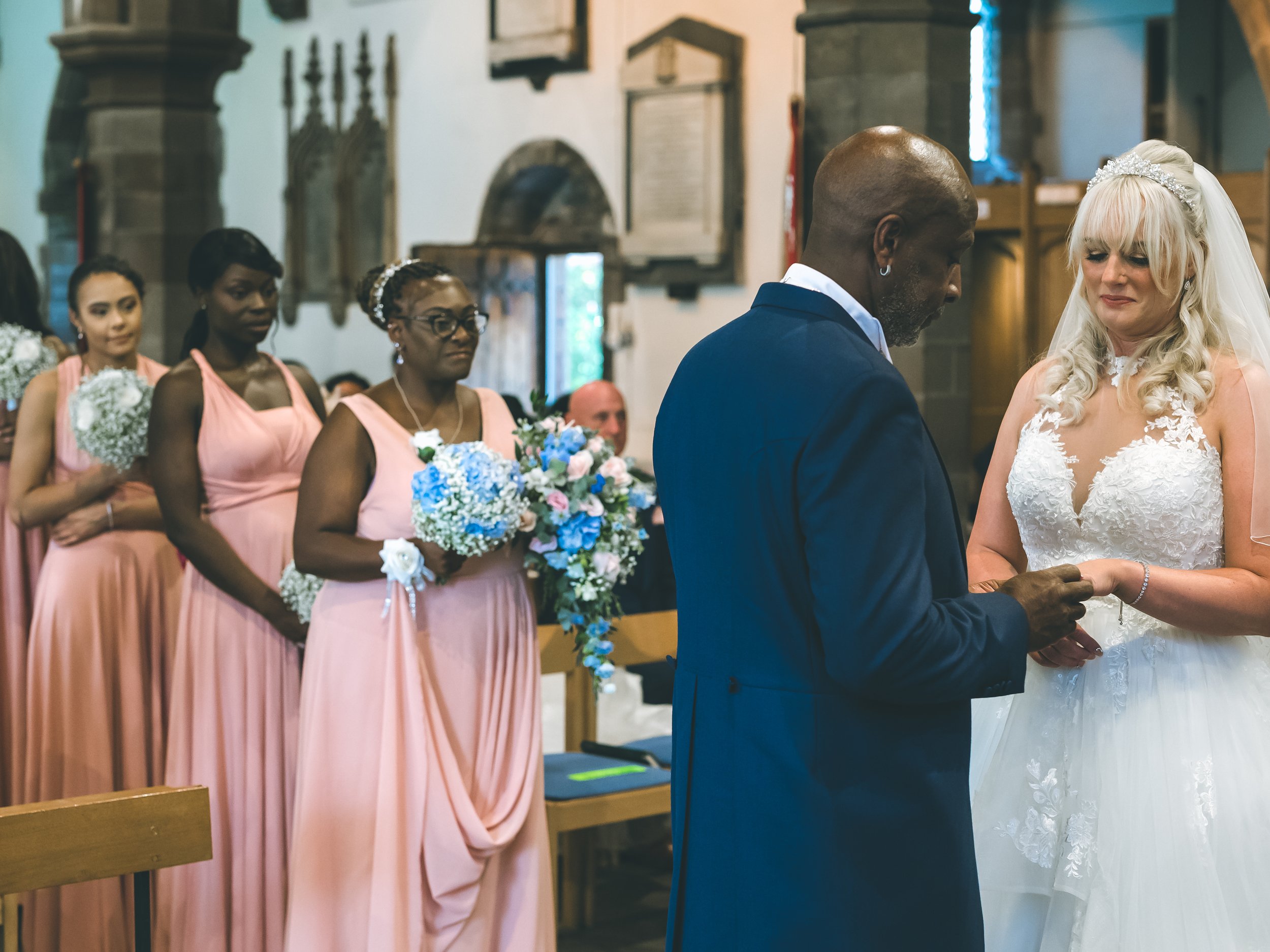 A bride and groom exchanging vows during a wedding ceremony in a church, with bridesmaids standing in the background holding bouquets.
