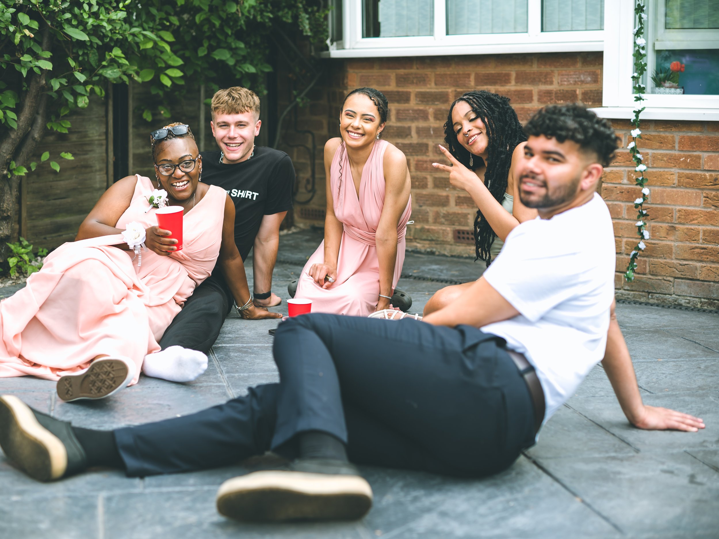 A group of five diverse young adults sitting on the ground in a backyard, smiling and enjoying each other's company, with a brick house and green bushes in the background.