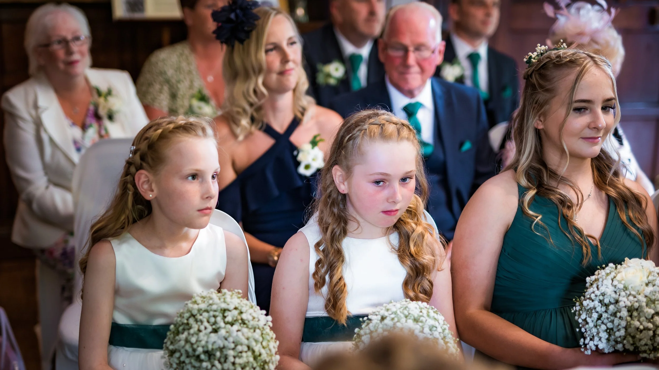 Young girls and women seated at a wedding ceremony, holding bouquets of white flowers, dressed in elegant dresses, with adults in formal attire behind them.