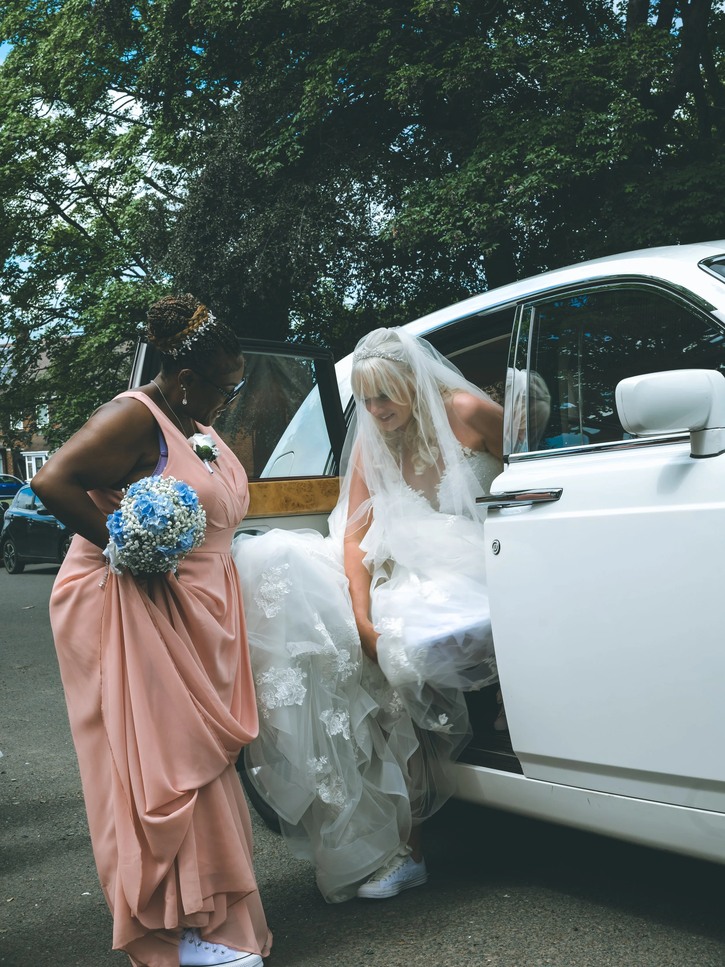 A bride in a wedding dress with a veil is stepping out of a white luxury car, assisted by a woman in a pink dress holding a bouquet of blue and white flowers. The scene is outdoors under a tree with green leaves.