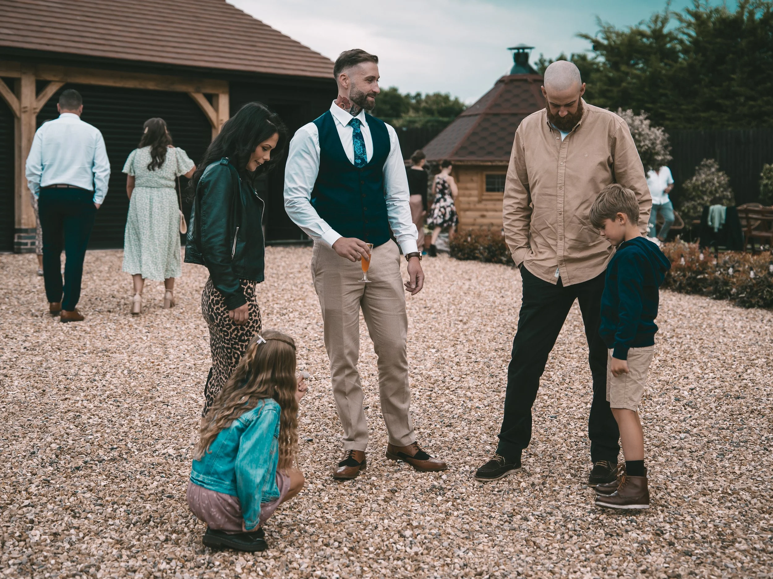 Group of people gathered outdoors at a social event, talking and socializing on a gravel pathway with a house and trees in the background.