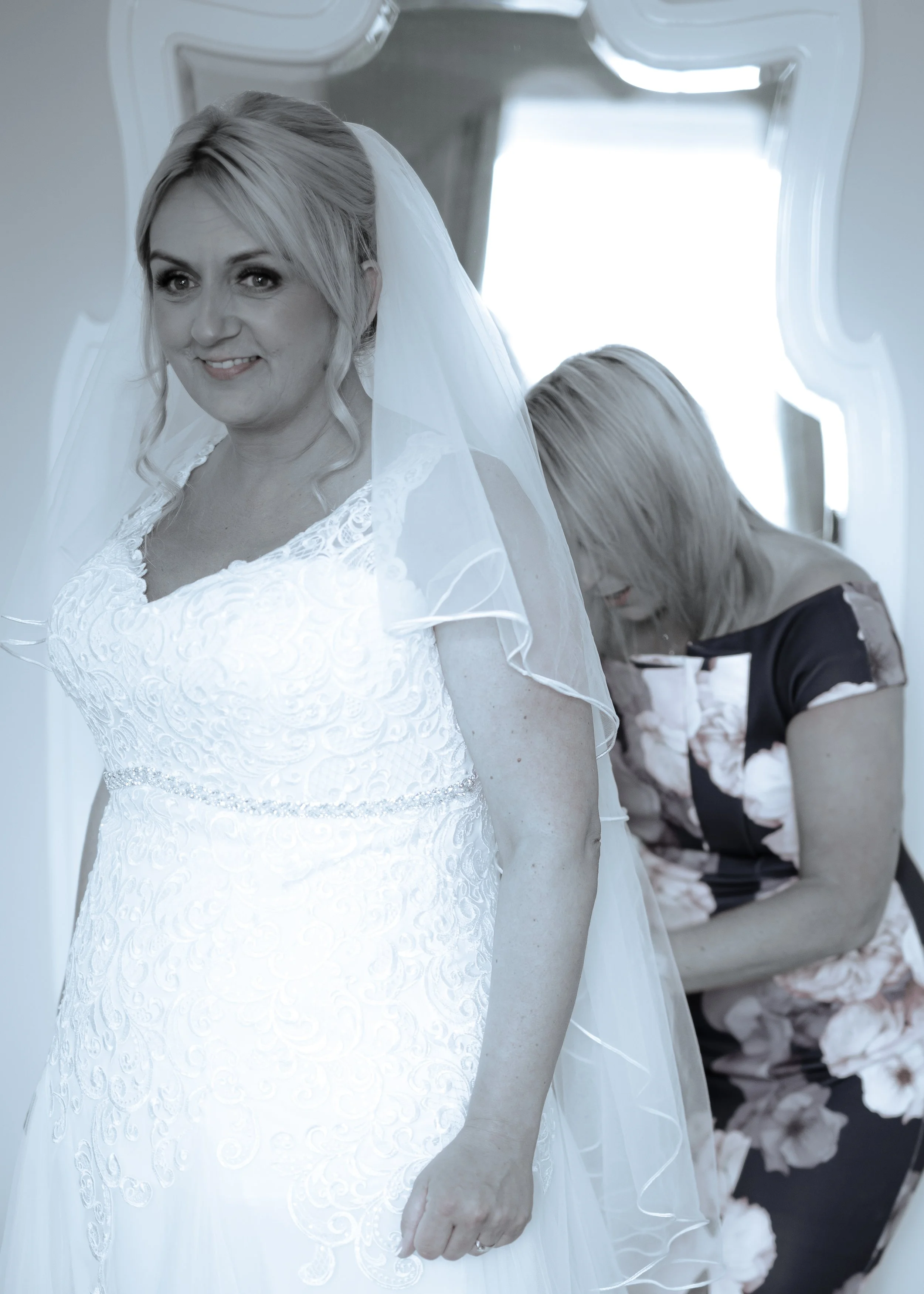 A bride in a white wedding dress with lace details, smiling while getting ready, as a woman adjusts her dress in the background.