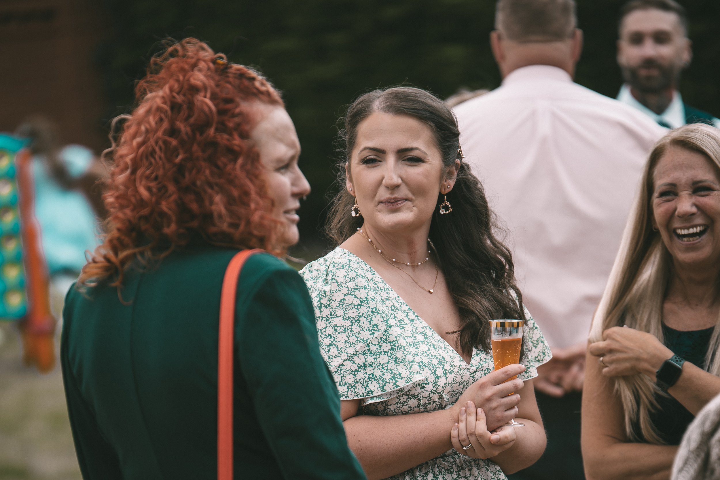 Group of people socializing outdoors, focusing on a woman with dark hair in a green patterned dress holding a glass of champagne, engaged in conversation.