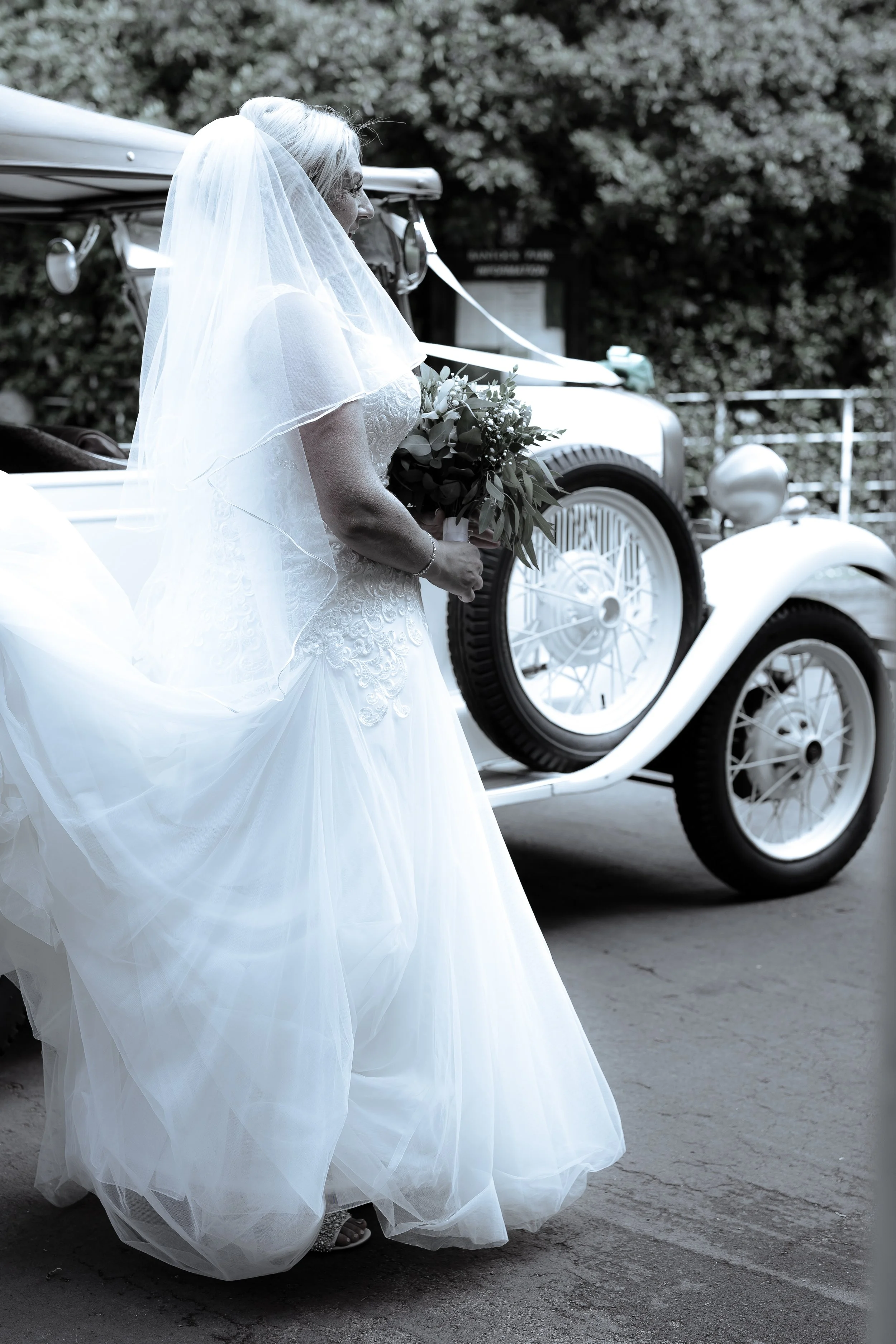 A bride in a white wedding dress holding a bouquet standing next to a vintage car.