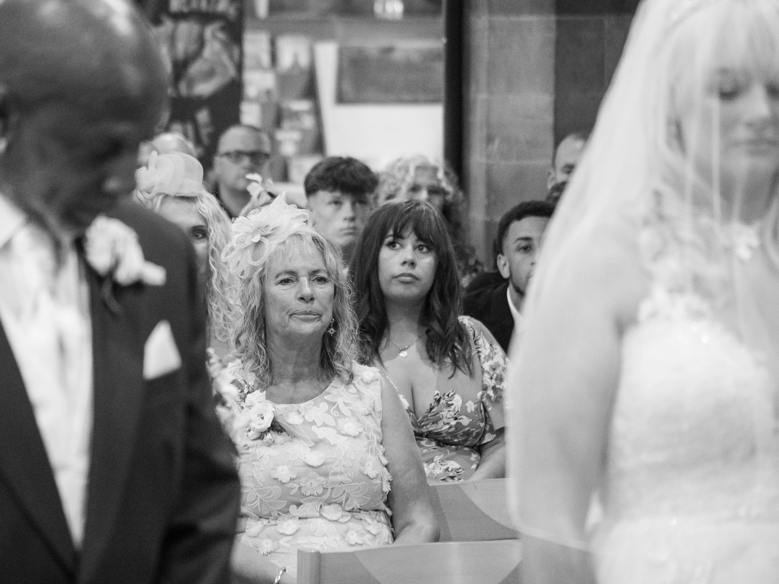 Black and white photo of people attending a wedding ceremony, with a woman wearing a floral dress and a fascinator, looking attentively, and a person dressed in a suit in the foreground.