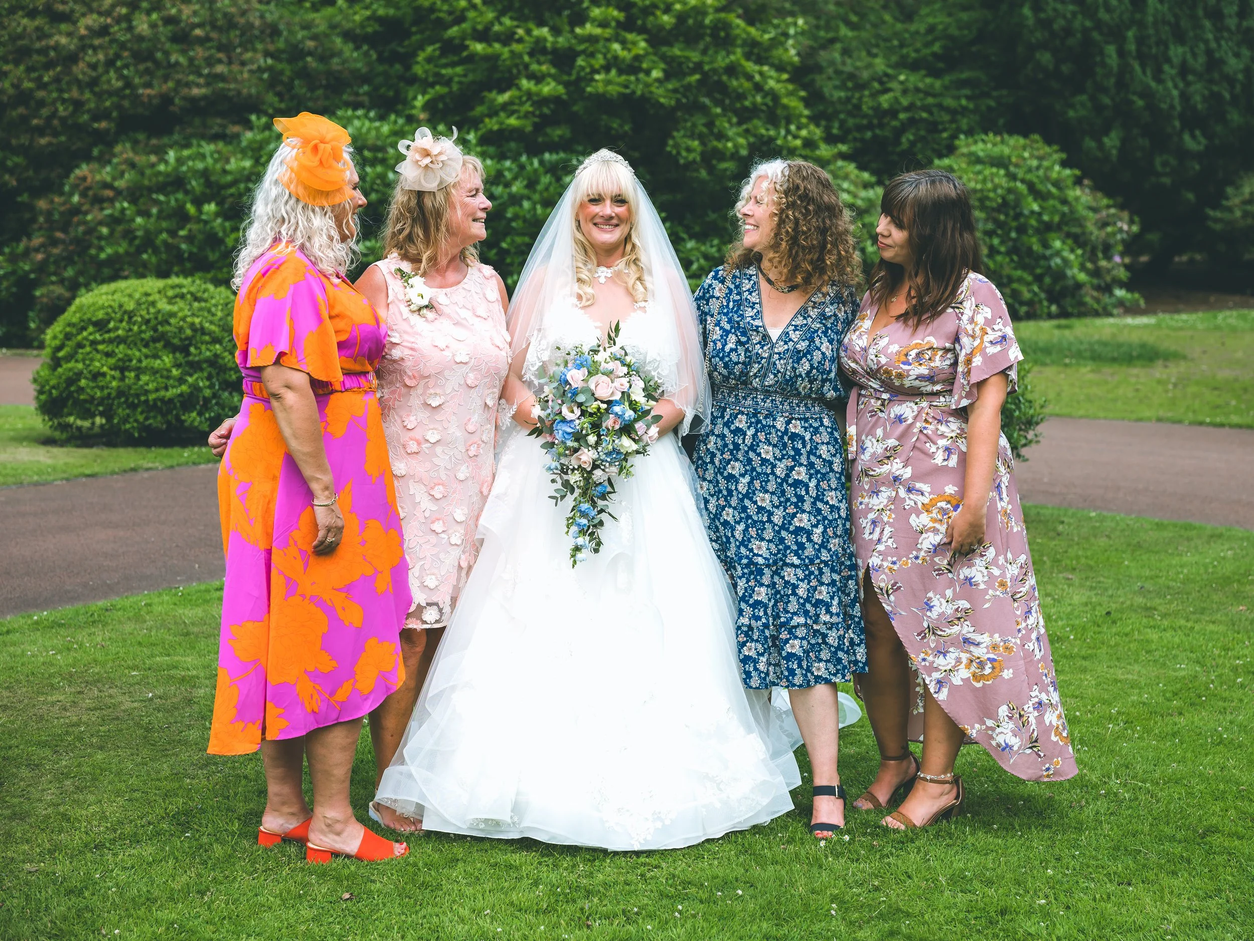A group of five women dressed in colorful dresses standing outdoors on grass, smiling and looking at each other, with green trees in the background. The woman in the center is wearing a white wedding dress and holding a bouquet, indicating it is a we
