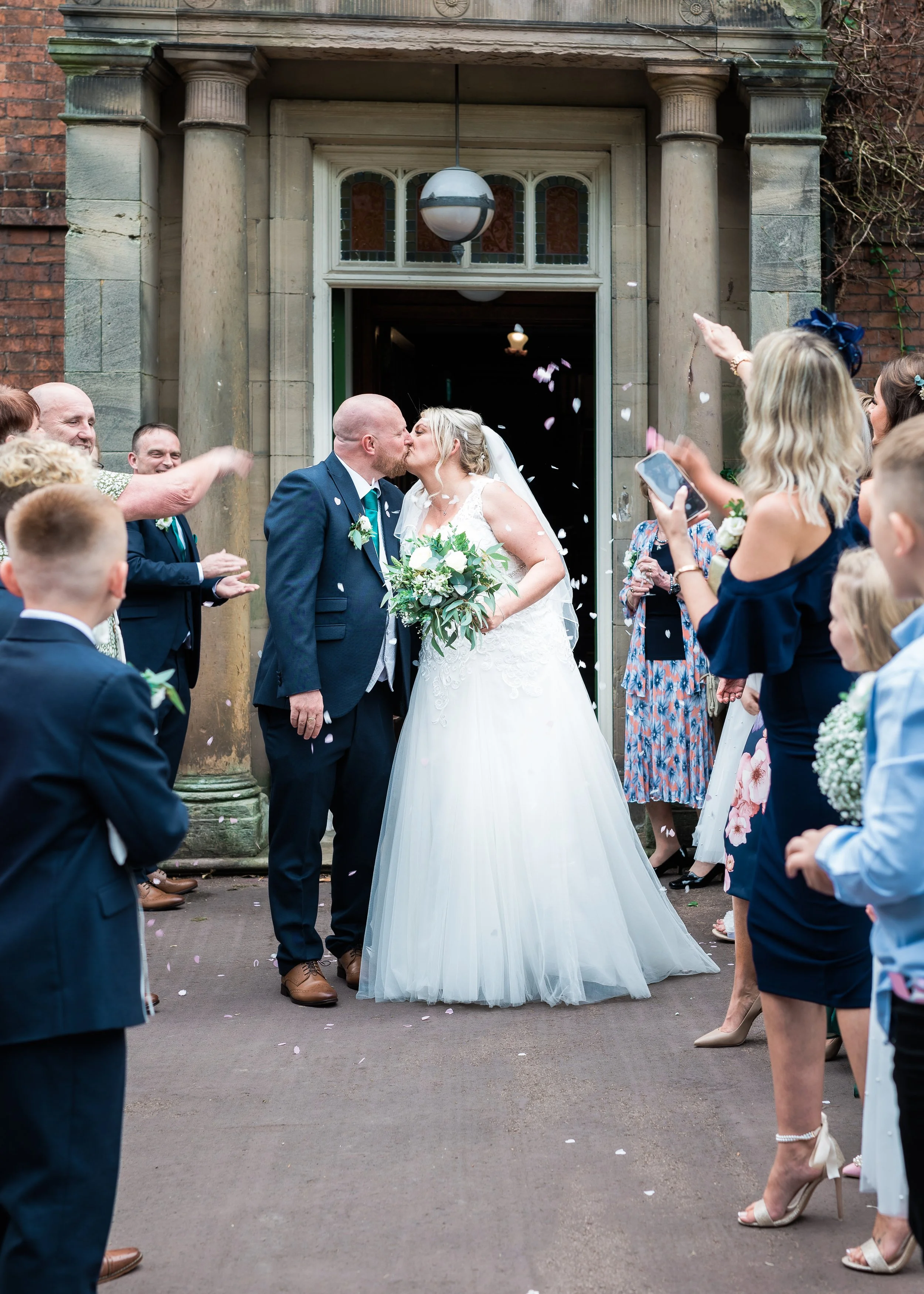 A wedding scene with a bride and groom sharing a kiss outside a building, surrounded by guests throwing flower petals and taking photos.