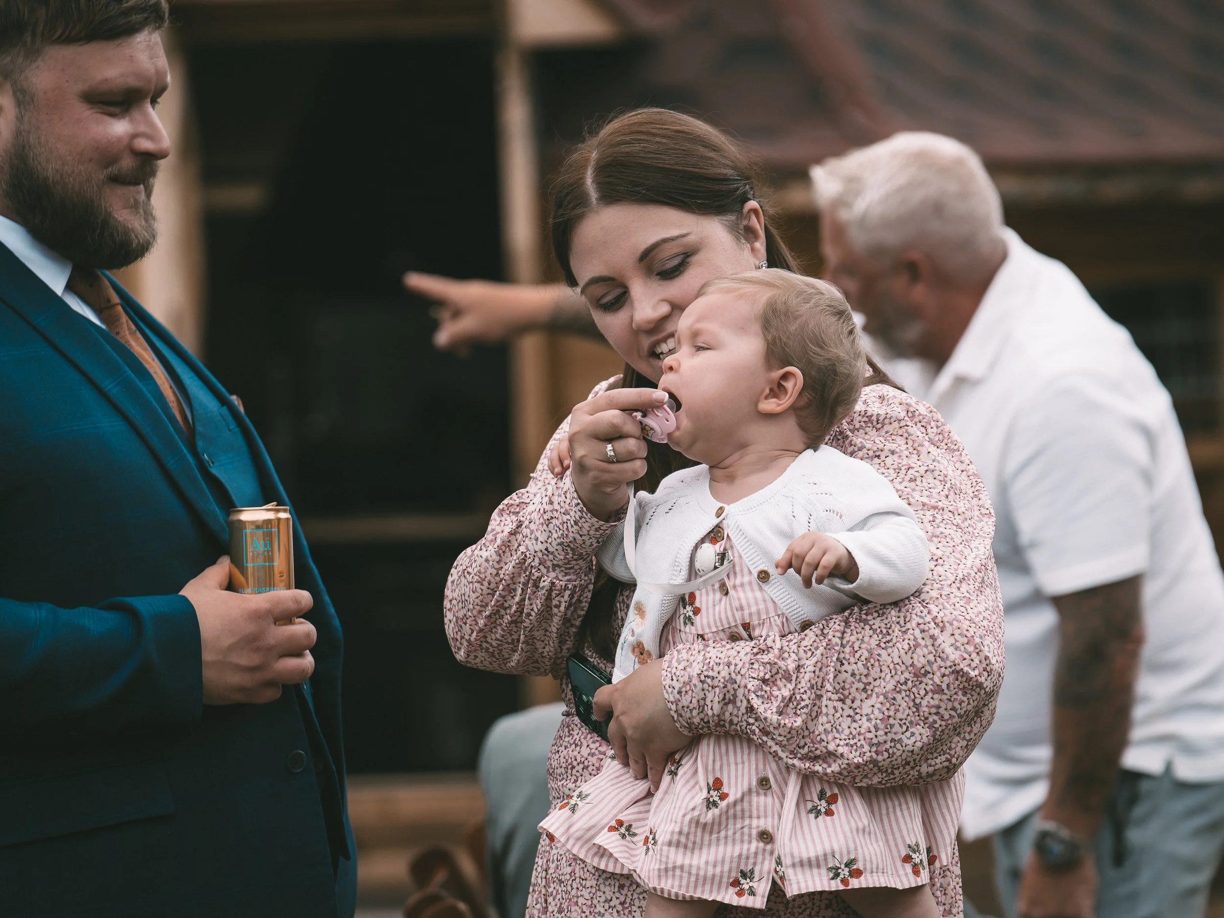 A woman is holding a young girl, who is yawning or talking, inside a gathering or event. The woman is smiling and has her arm around the girl. There are two men visible in the background, one in a white shirt and the other in a blue suit and tie, eng
