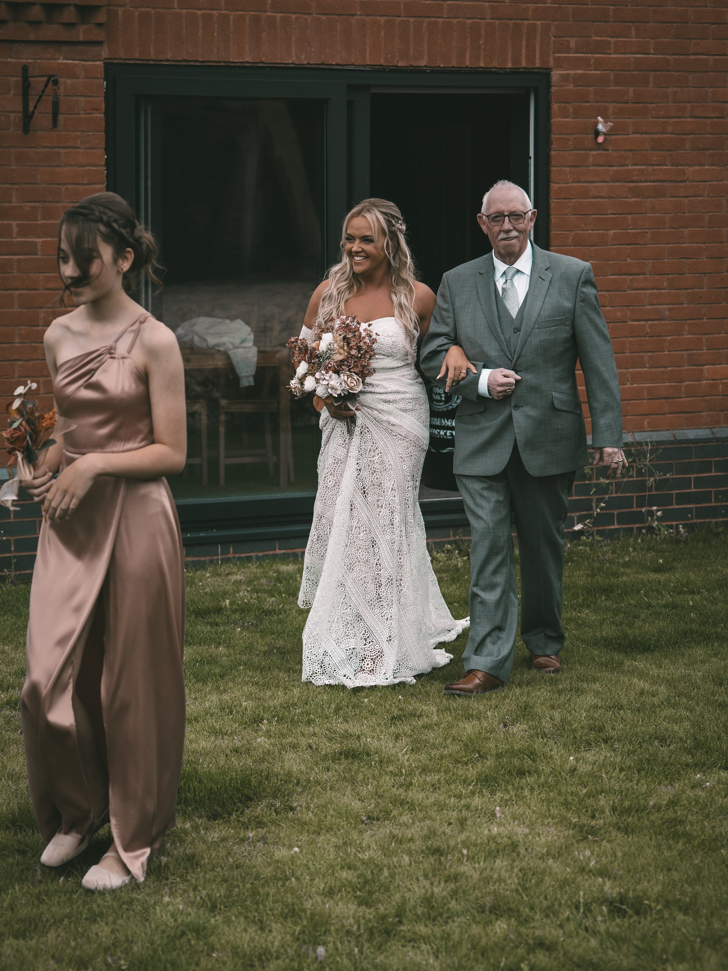 A woman in a white lace wedding dress holding a bouquet, walking arm-in-arm with an elderly man in a gray suit, outside a brick building with a large glass door.