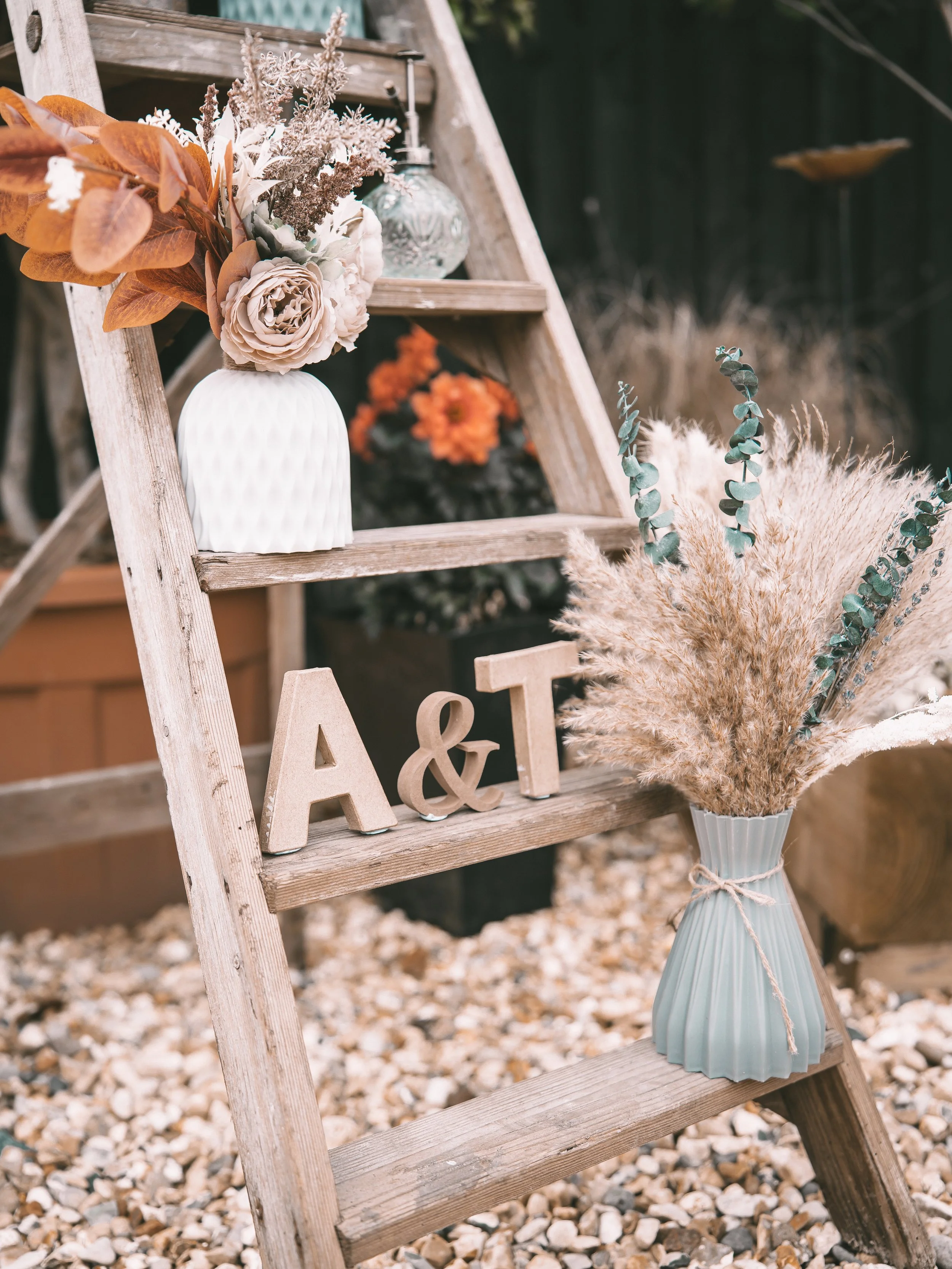 A rustic wedding or event decoration setup with a wooden ladder, floral arrangements, and decorative letters on the bottom step. The setup includes a vase with pampas grass and eucalyptus, a bouquet with dried and artificial flowers, and a glass jar 