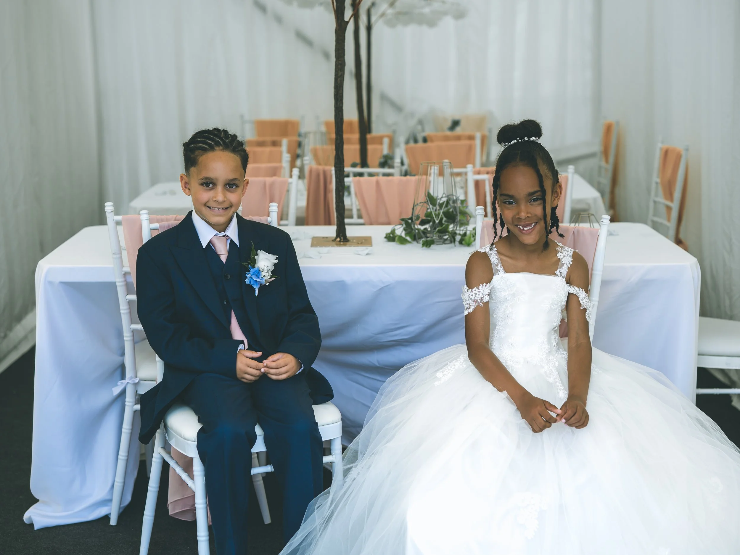 Two children dressed in formal attire, a boy in a navy suit and a girl in a white dress, sitting at a decorated table in a banquet hall.