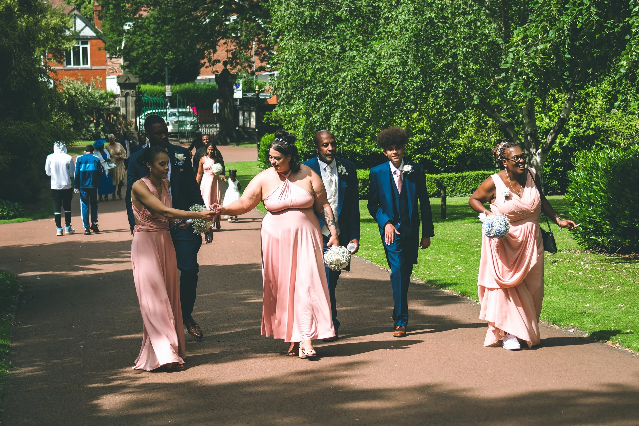 A diverse group of people dressed in formal attire walking in a garden or park during a wedding celebration. Women in pink dresses hold bouquets, while men in suits walk beside them.
