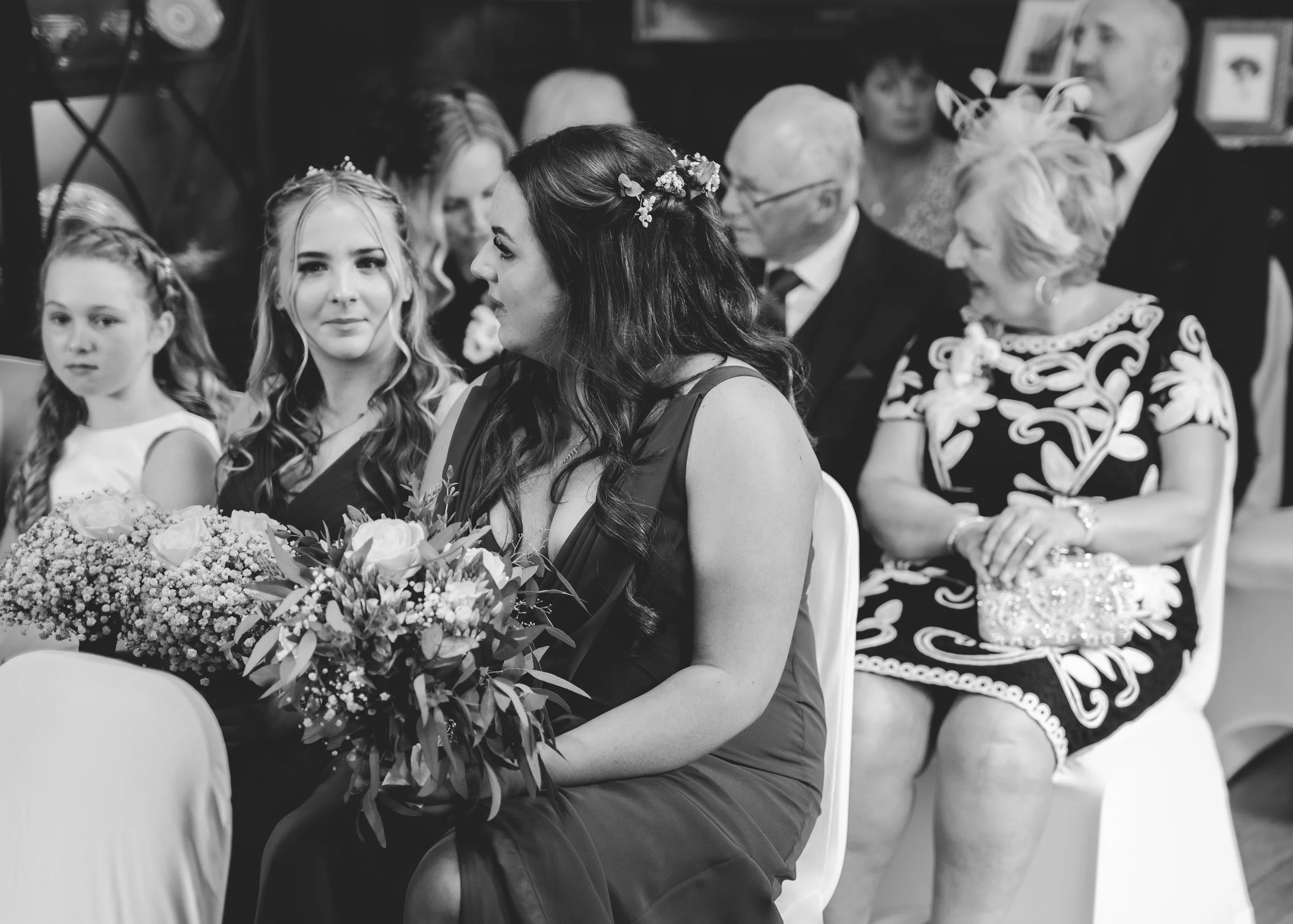 Black and white photo of a woman with flowers in her hair, sitting at a formal event beside a bouquet of flowers, with children and other adults in the background.