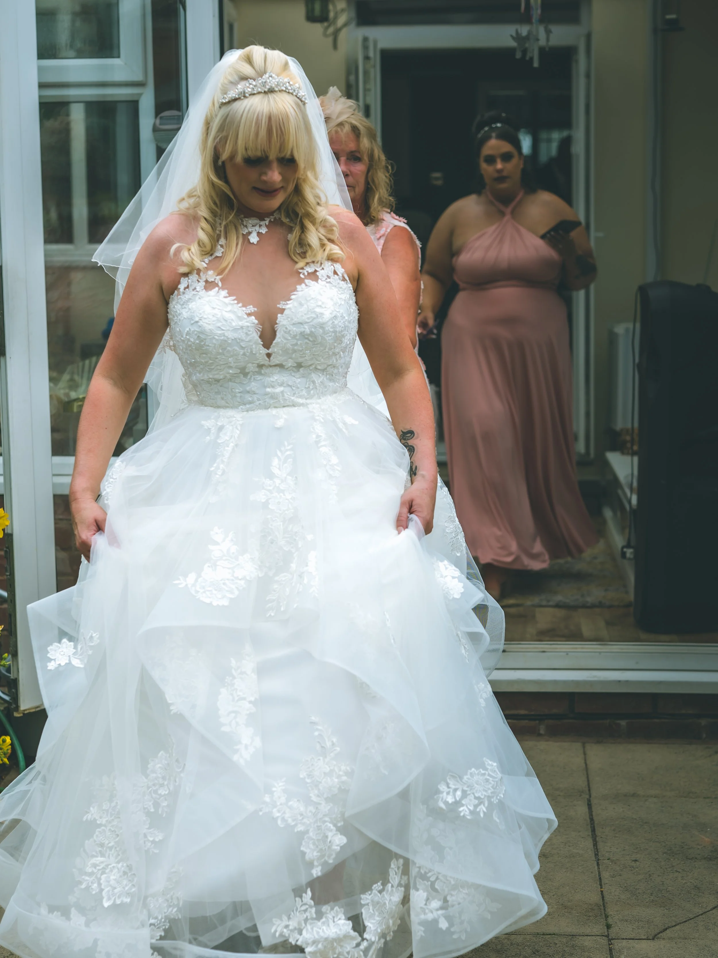 A bride in a white wedding dress with lace details and a veil, holding the skirt of her dress, standing outside near a glass door. Behind her, there are two women, one with blonde hair and another with dark hair, both dressed in pastel-colored dresse