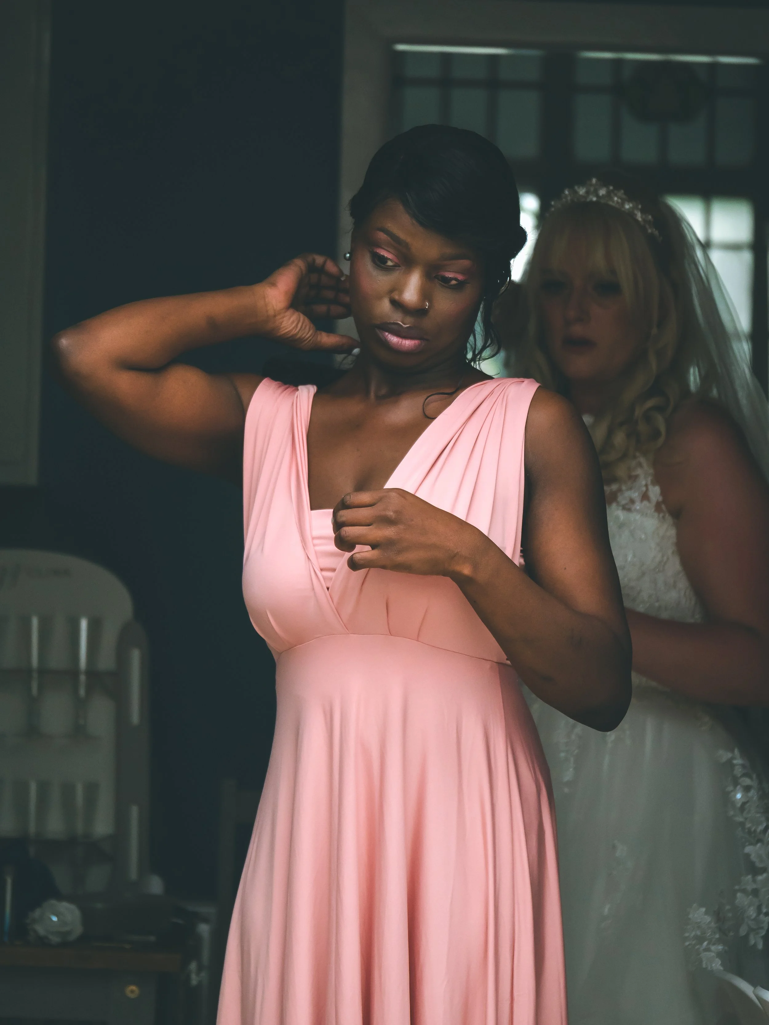 A woman in a pink dress trying on a dress in a fitting room while another woman in a wedding dress looks on in the background.