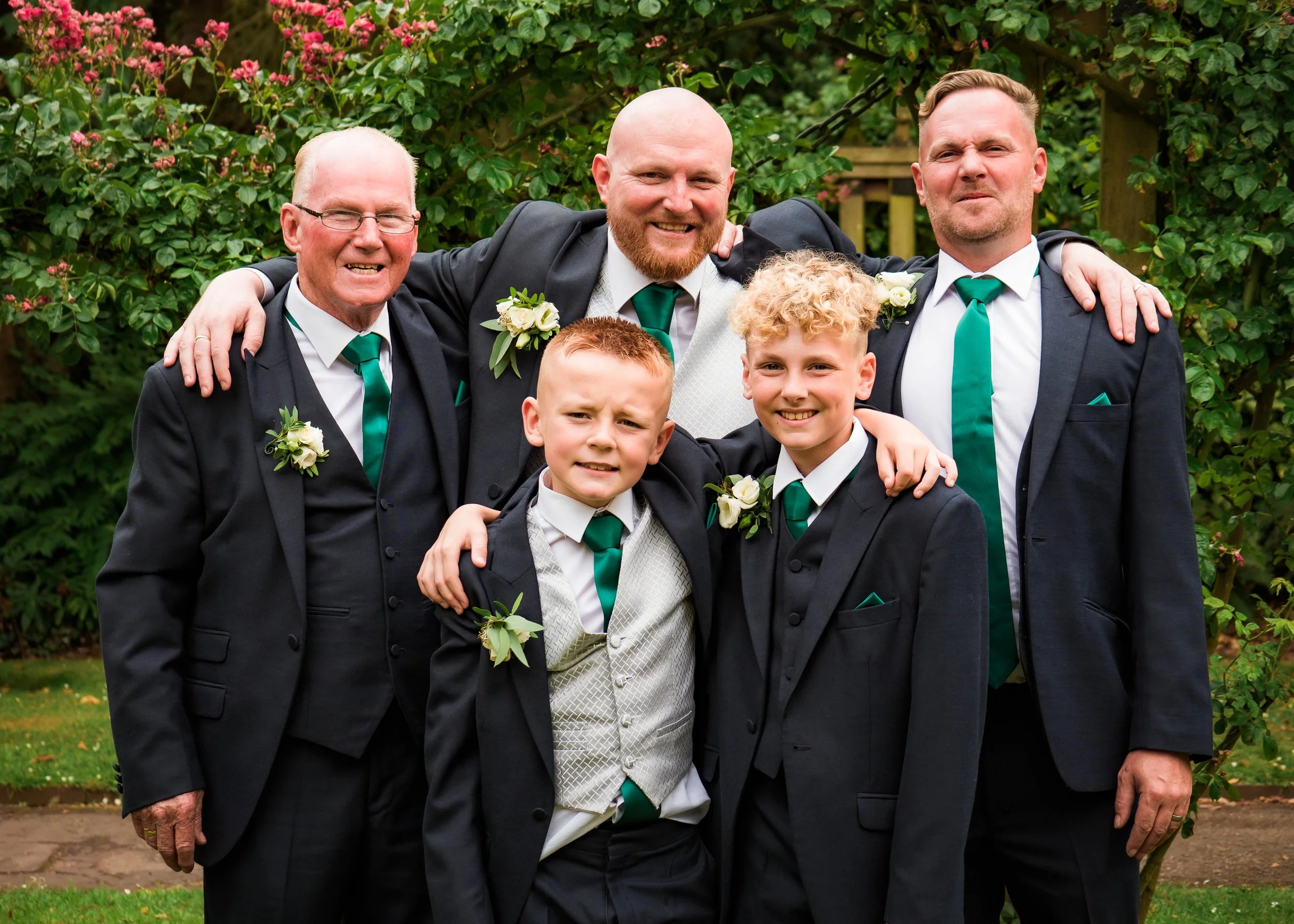 Four men and two boys dressed in formal tuxedos with green ties and white flowers on their jackets, standing outdoors in front of bushes with pink flowers, smiling and posing for a group photo.