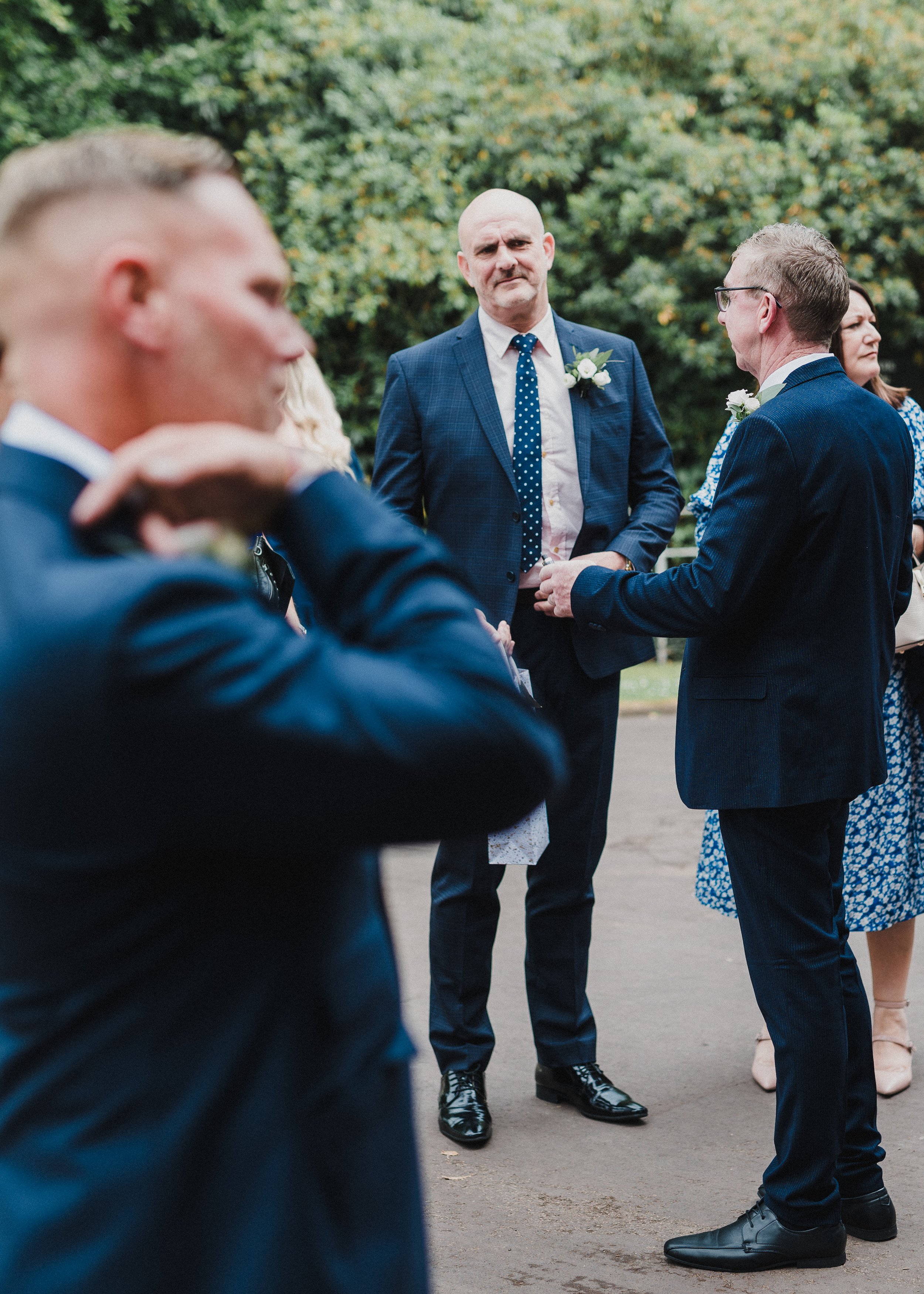 A group of people dressed in formal attire, including suits and dresses, are gathered outdoors, suggesting a wedding or special event. The focus is on a man with a bald head and a serious expression, wearing a blue checked suit and a polka-dot tie, s