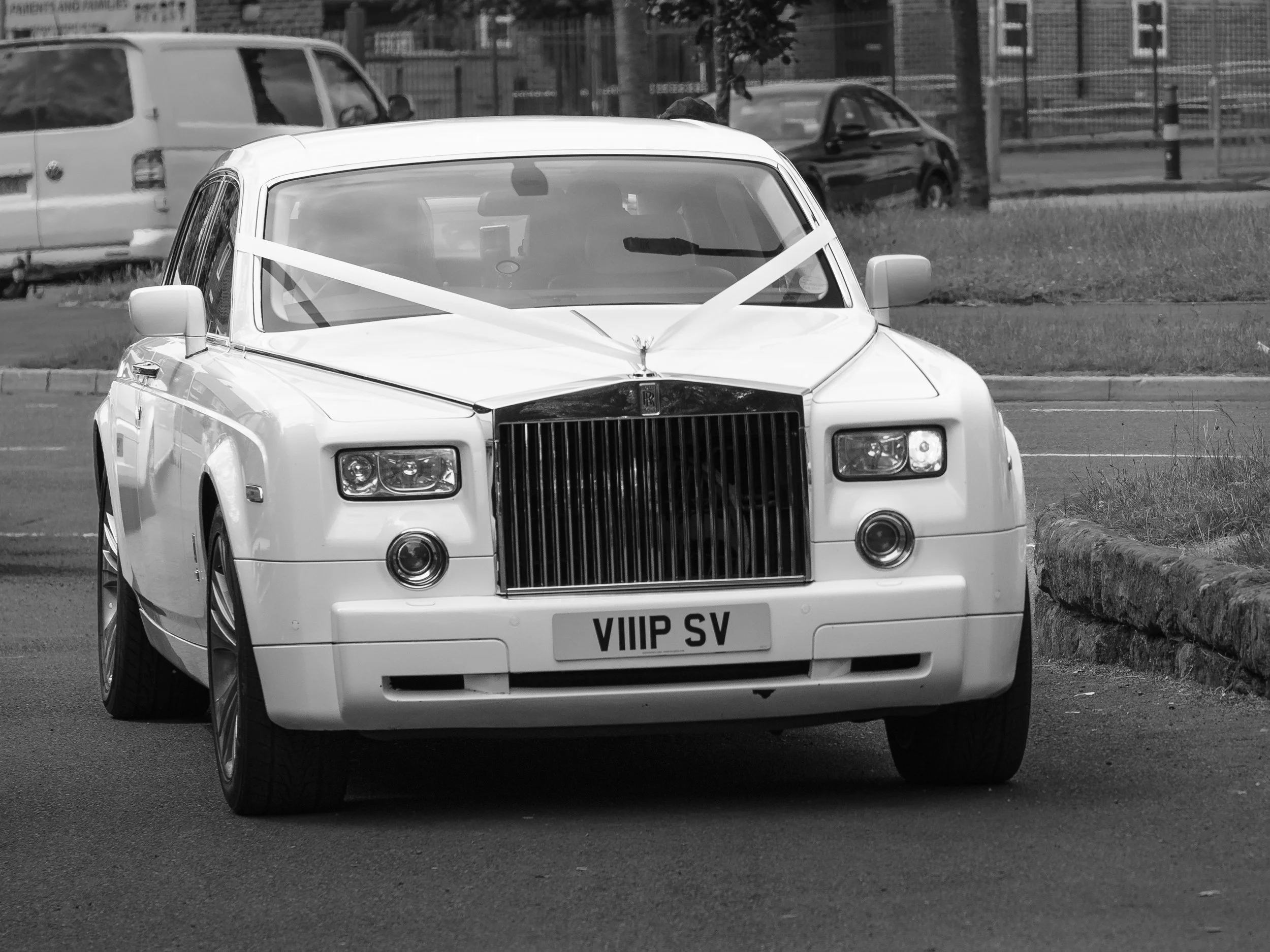 White luxury car with a prominent front grille, parked in a parking lot with other vehicles and a fence in the background, decorated with ribbons tied to the hood and windshield.