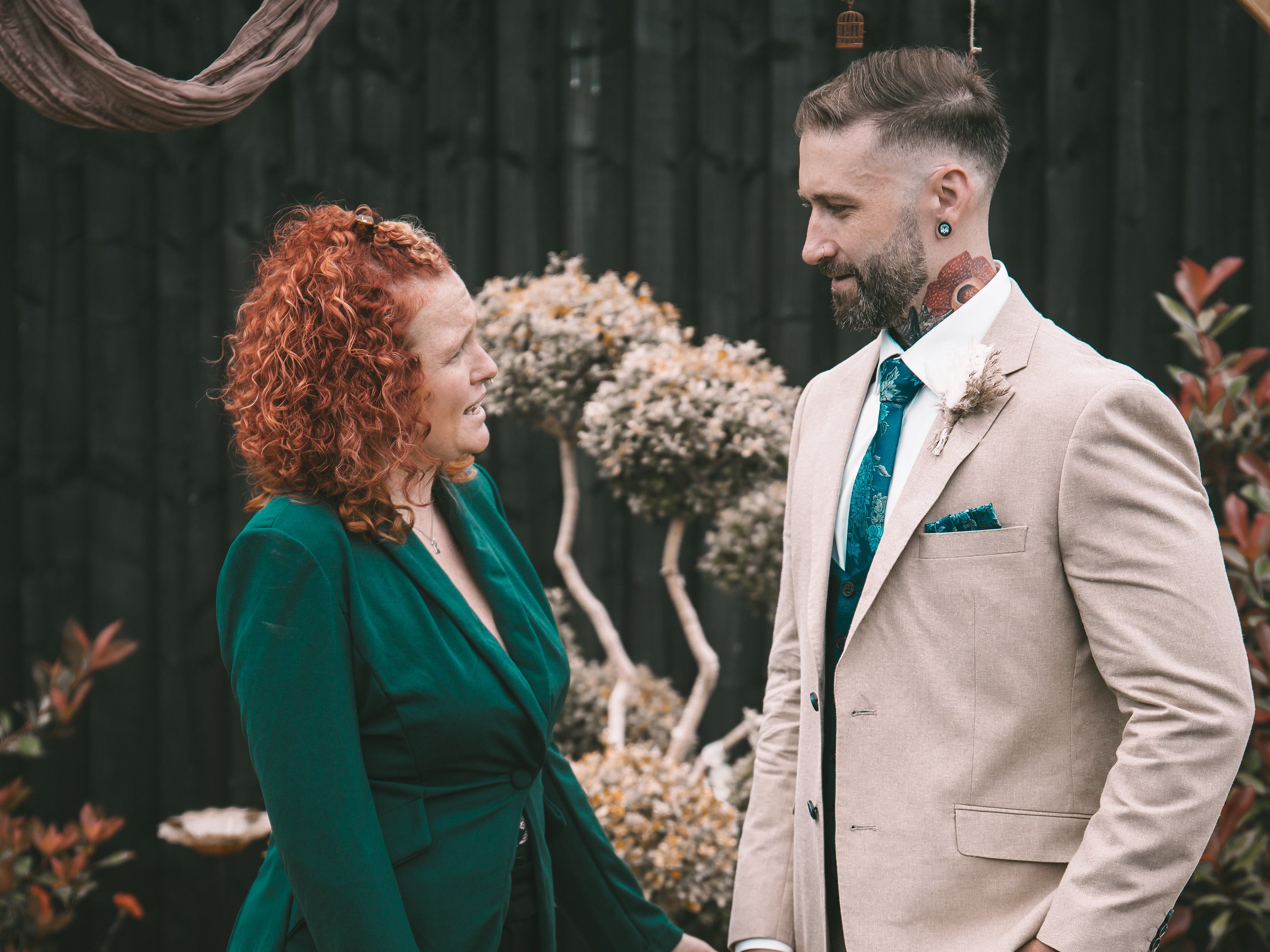 A woman and a man, both with bright red hair, facing each other and smiling outdoors. The woman is wearing a green blazer and the man is wearing a beige suit with a teal tie, a floral pocket square, and has tattoos and earrings. In the background, th