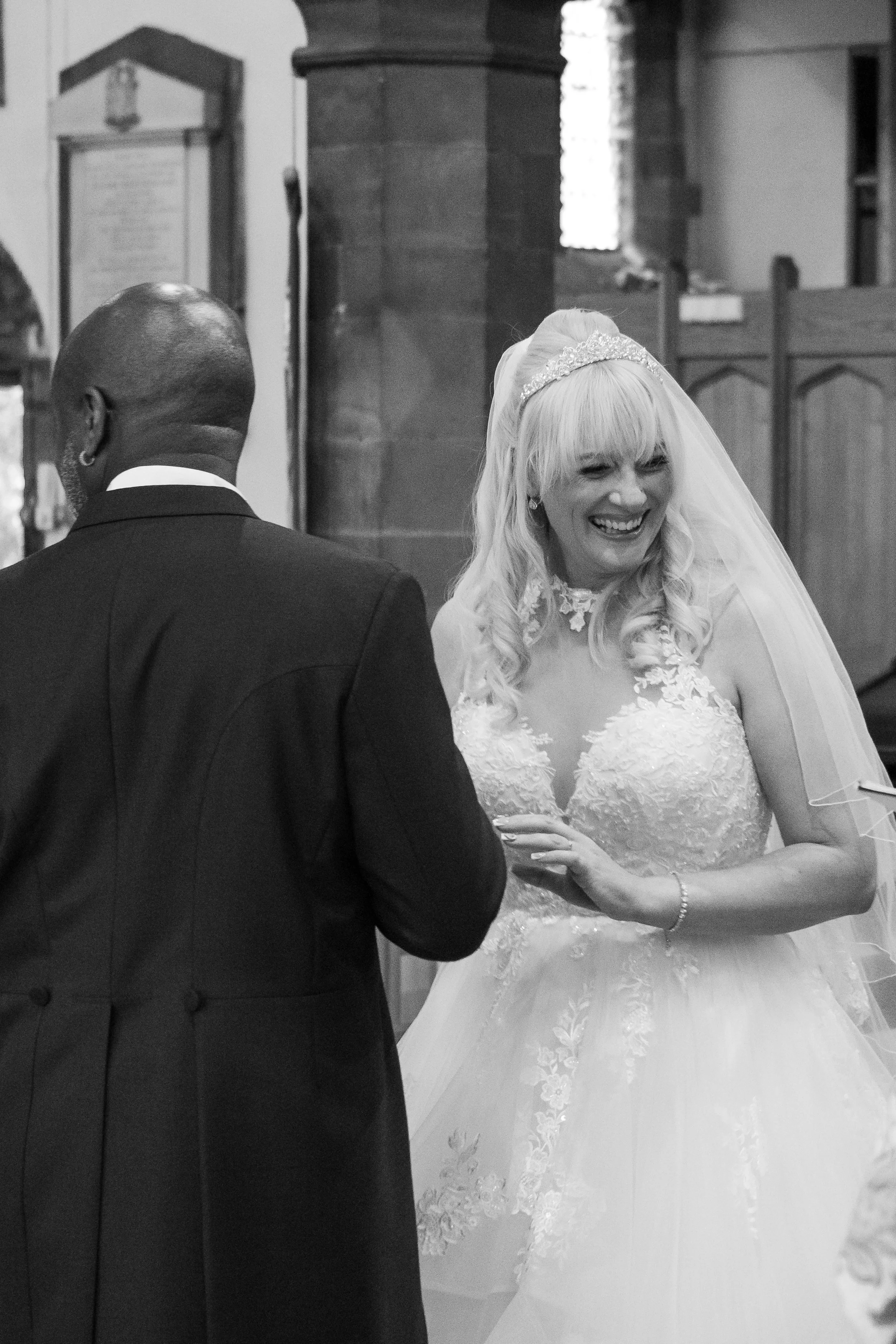 A black and white photo of a bride and groom during their wedding ceremony inside a church. The bride, smiling and wearing a lace wedding gown and a veil, is holding the groom's hand. The groom, seen from behind, is dressed in a dark suit.