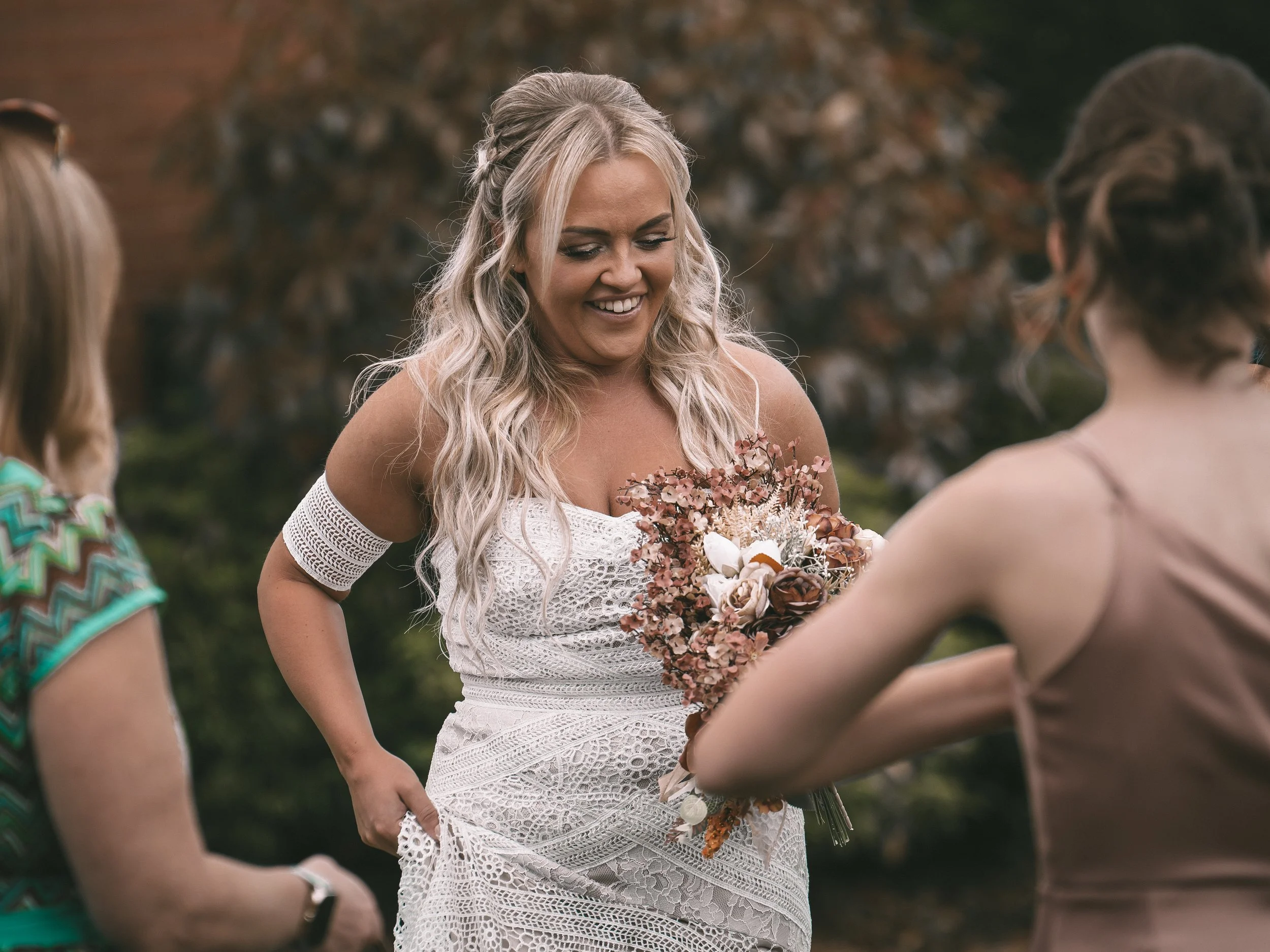 A woman with long blonde hair styled in loose curls, wearing a white lace dress and holding a bouquet of pink, peach, and white flowers, is smiling and talking to other women outdoors during daytime.