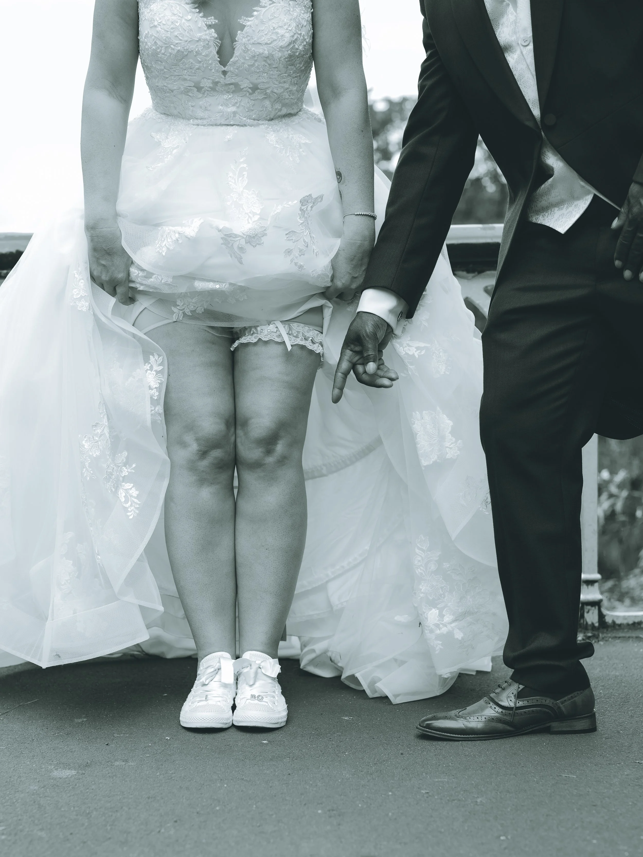 A bride and groom holding hands, with the bride lifting her wedding dress slightly to show her legs and shoes, standing outdoors.