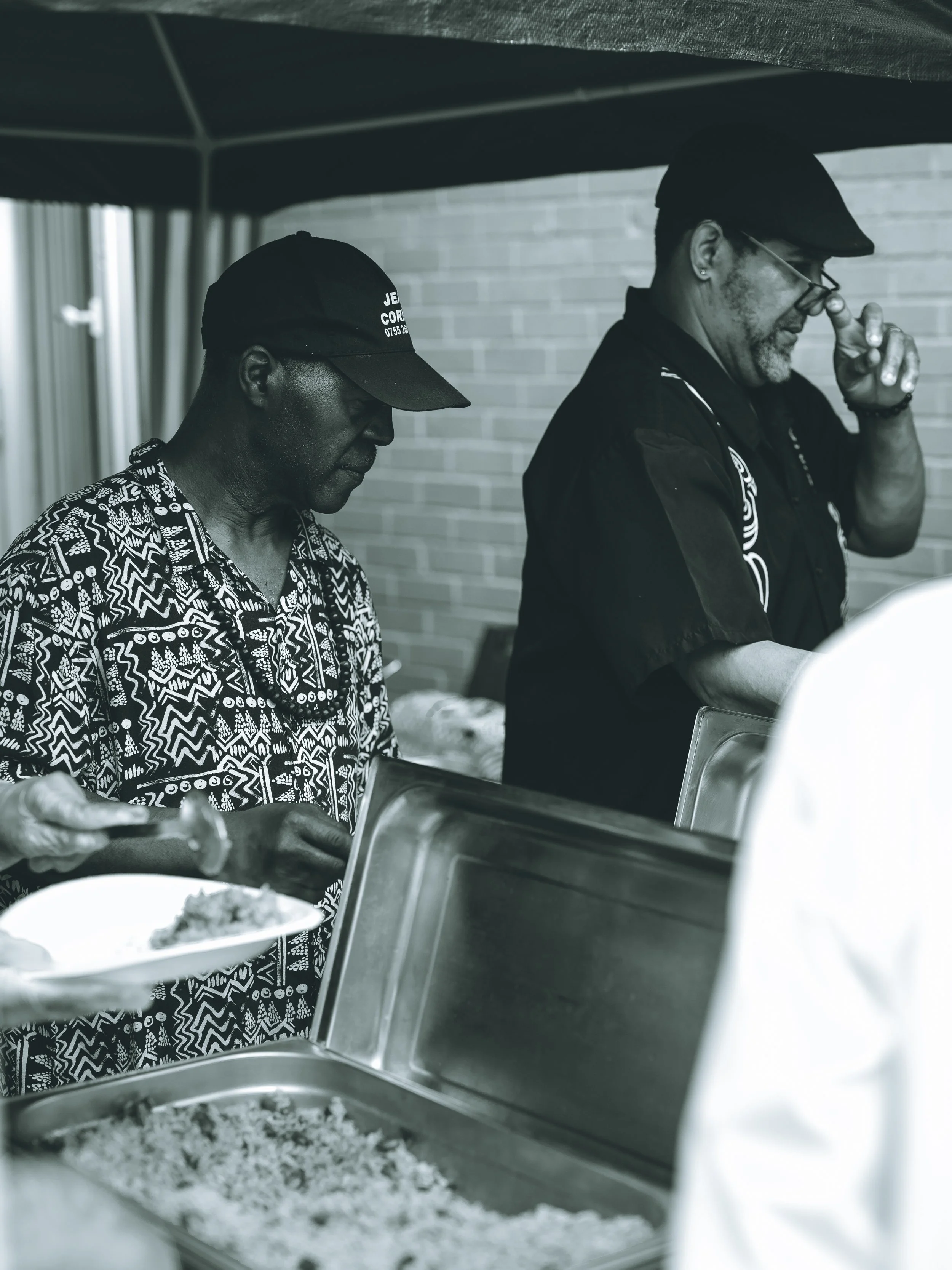 Two men standing near a food serving table, with one serving food from a tray. The man on the left is wearing a patterned shirt and a cap, and the man on the right is wearing glasses, a cap, and a dark jacket. They are under a canopy, with a brick wa