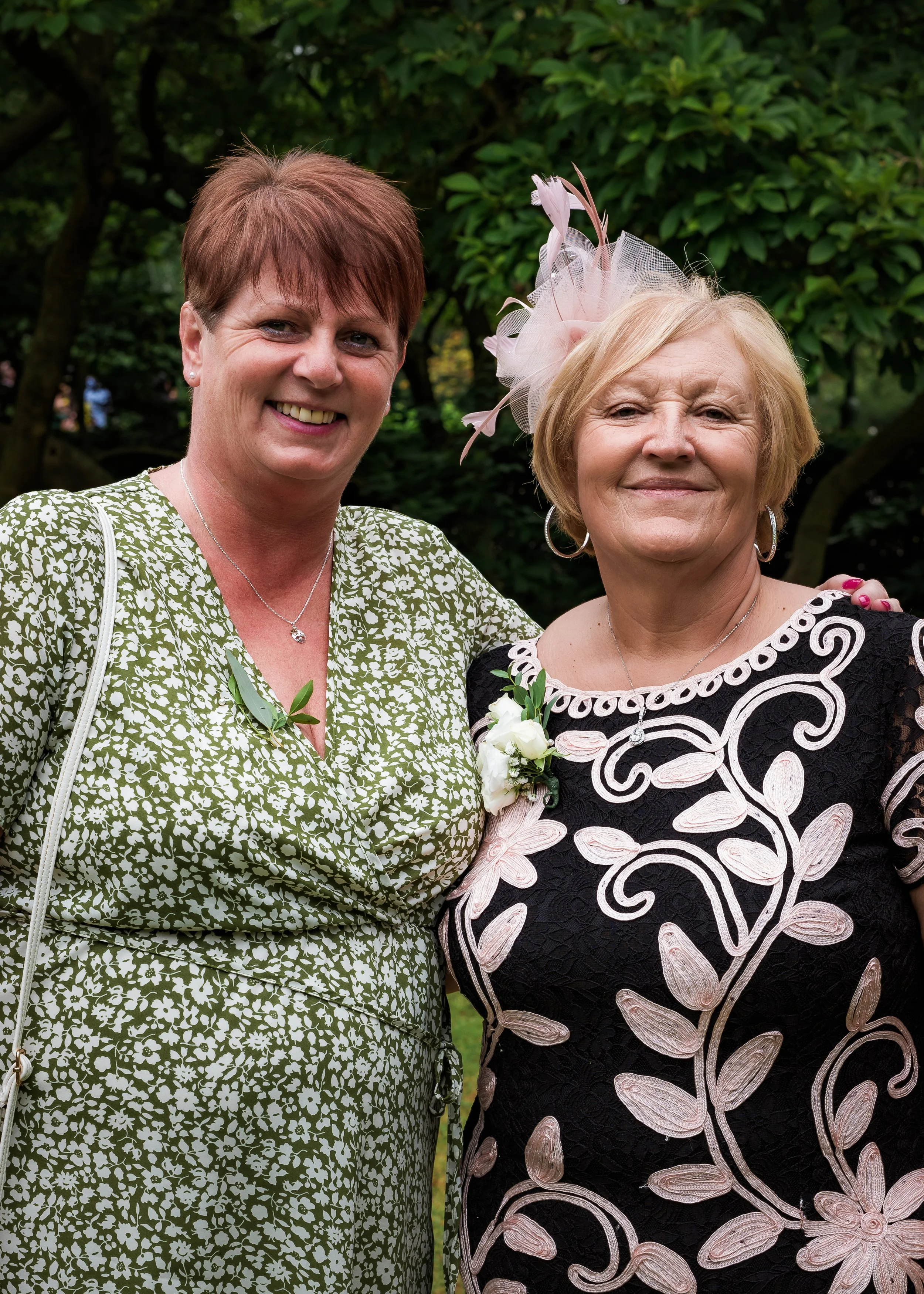Two women smiling and posing together outdoors with greenery in the background. One woman has short red hair and is wearing a green and white patterned dress, the other has light blonde hair, is wearing a black dress with floral embroidery, and a pin