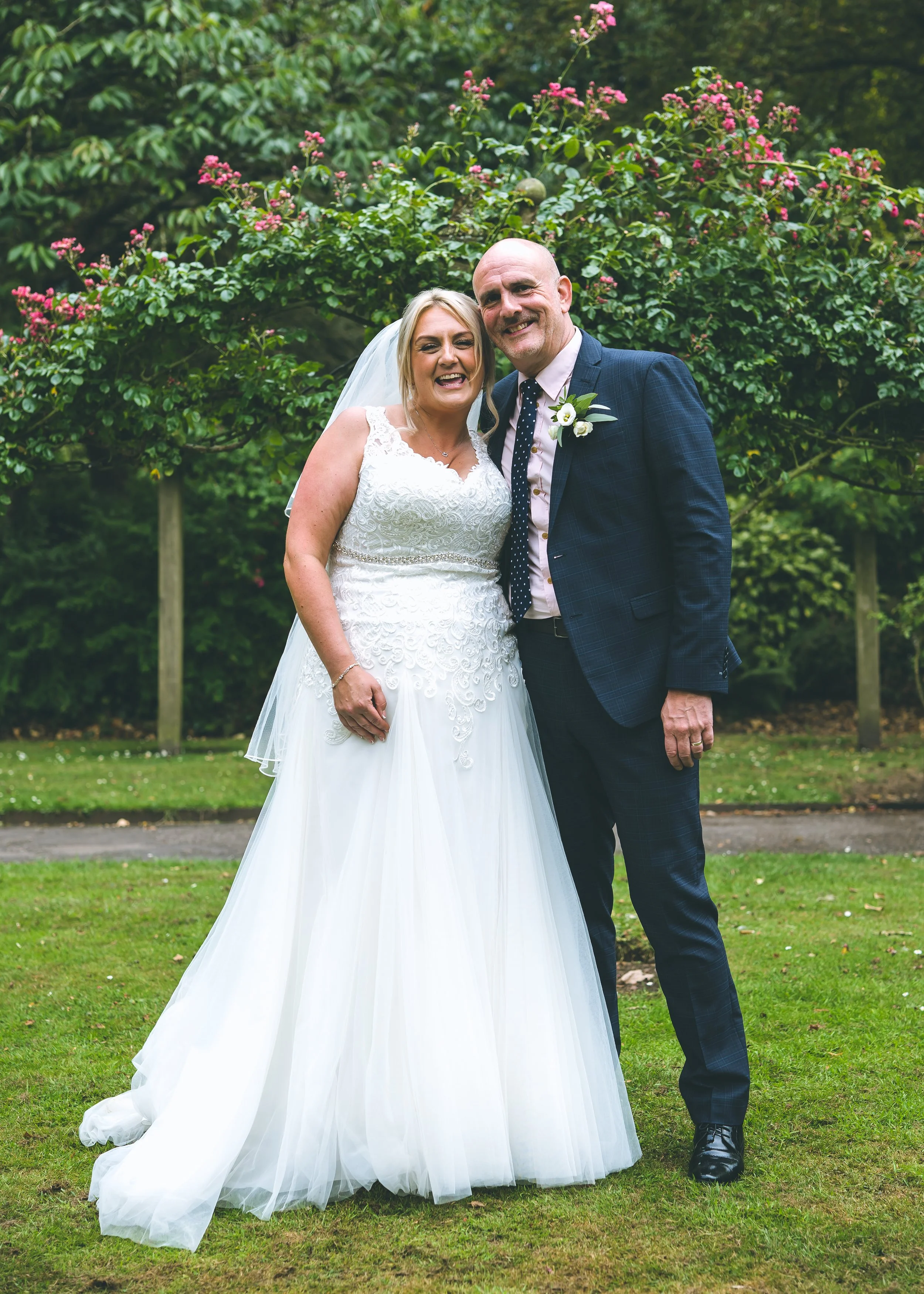 A bride in a white wedding gown and veil standing next to a groom in a dark suit with a checkered pattern, both smiling outdoors in front of a bush with pink flowers and green trees.