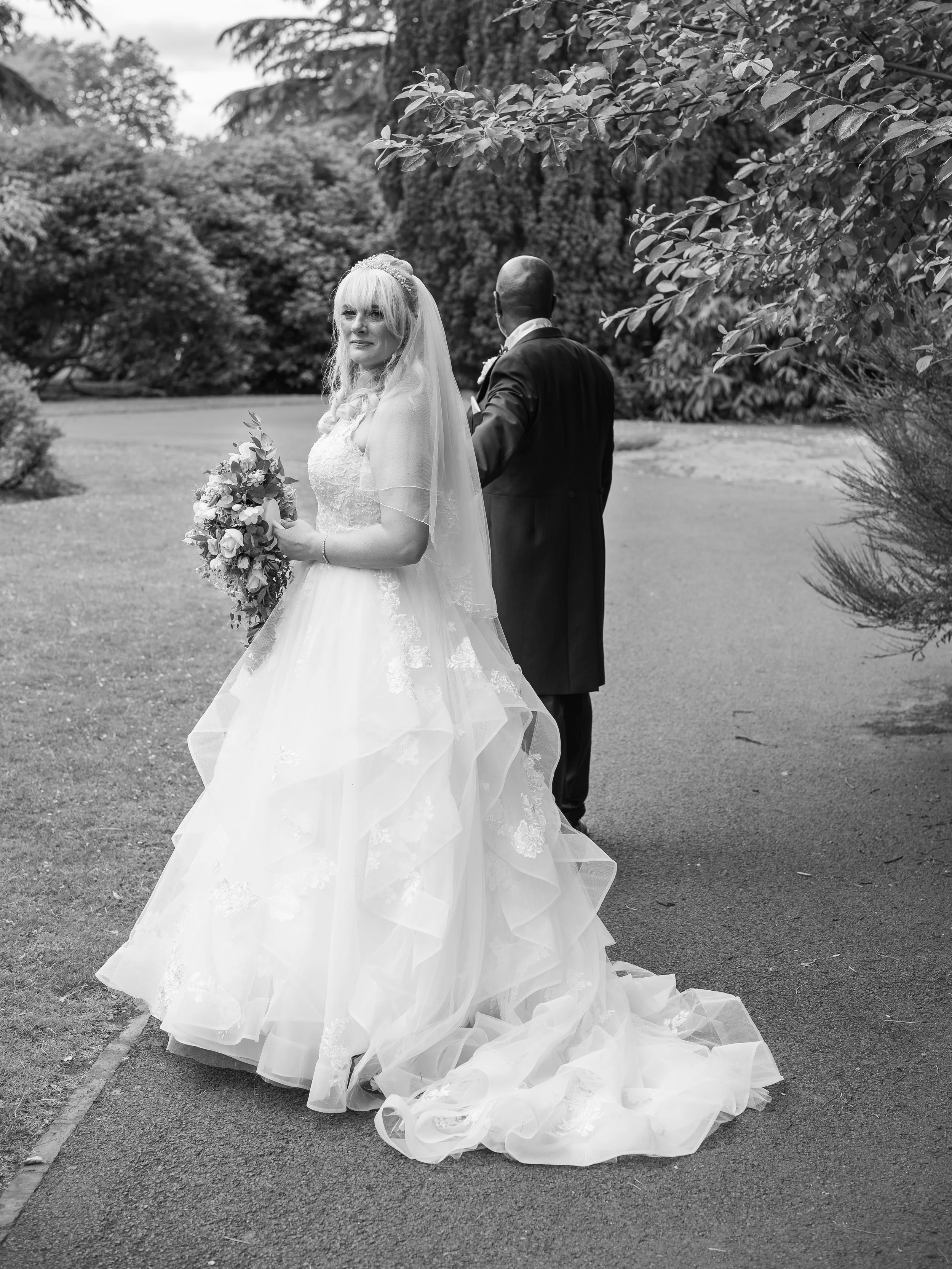 A bride in a wedding dress holding a bouquet, standing on a paved path outdoors with trees in the background, with a man behind her.